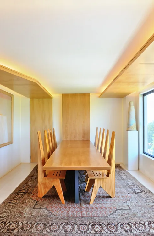A long wooden dining table is surrounded by angular, high-back wooden chairs on an ornate, patterned area rug under warm cove lighting.