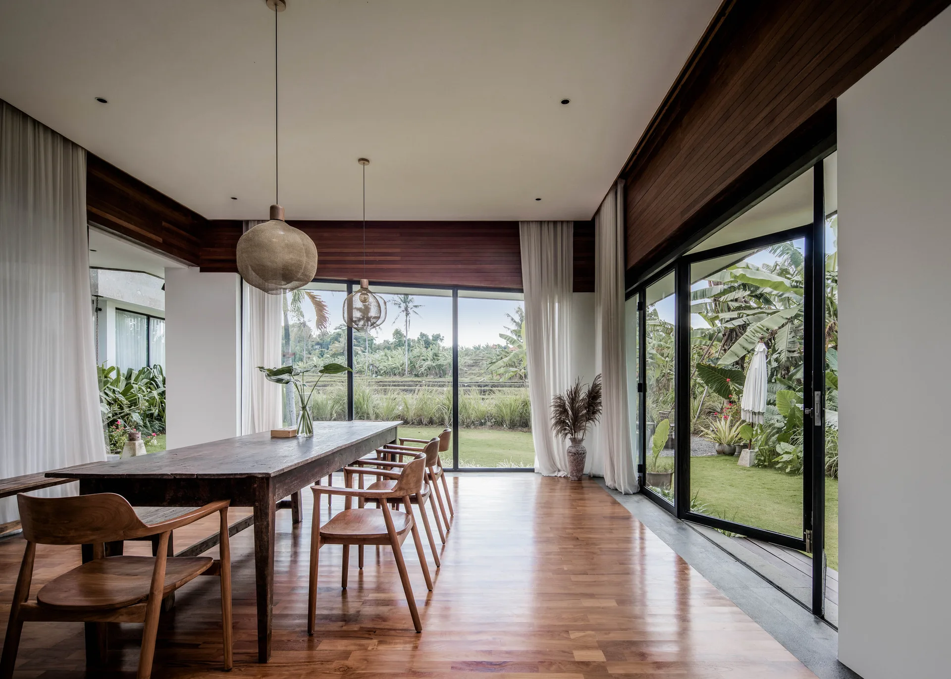A dining room features a long dark wood table, wooden chairs, woven pendant lights, and sliding glass doors opening to a lush tropical garden.