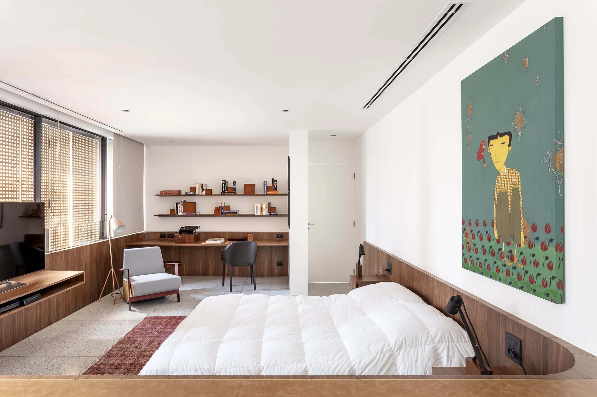 A modern bedroom features a white bed with a long wooden headboard, a built-in study desk with floating shelves, and a large green portrait painting.