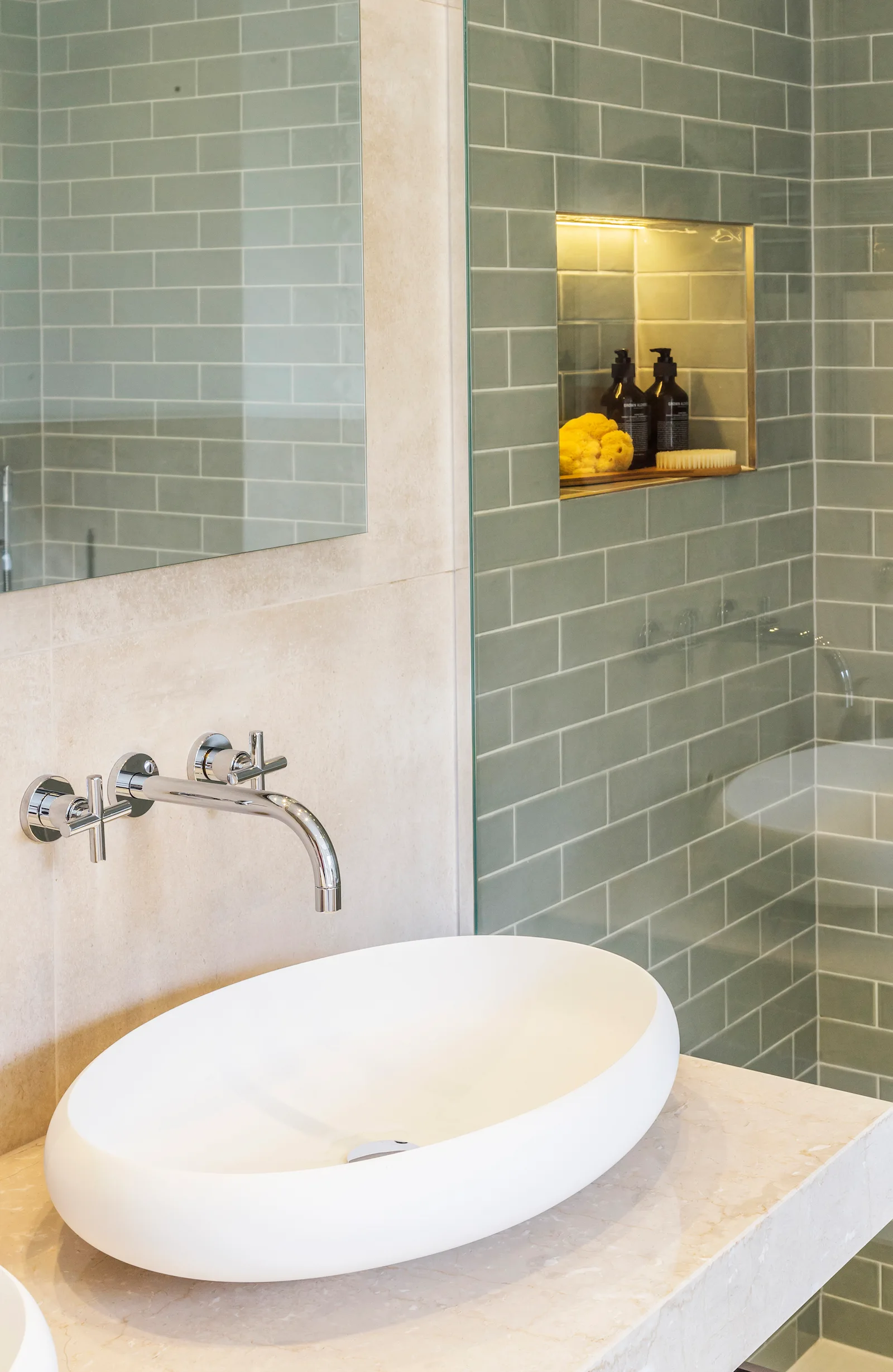 A modern white vessel sink sits on a beige stone vanity next to a glass shower partition revealing glossy sage green subway tiles.