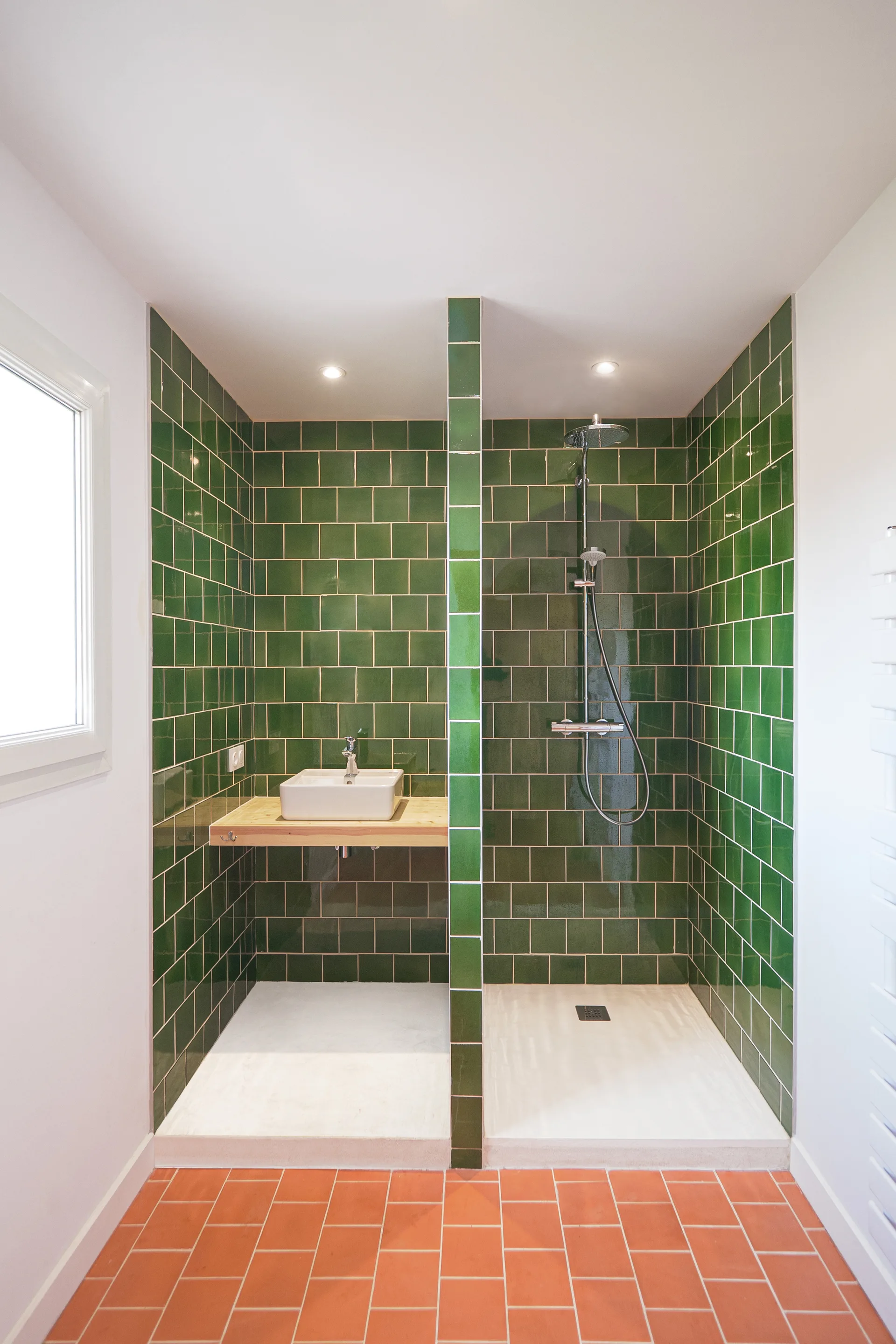 A symmetrical modern bathroom features two green tiled stalls divided by a central partition, one with a white sink and the other with a chrome shower.
