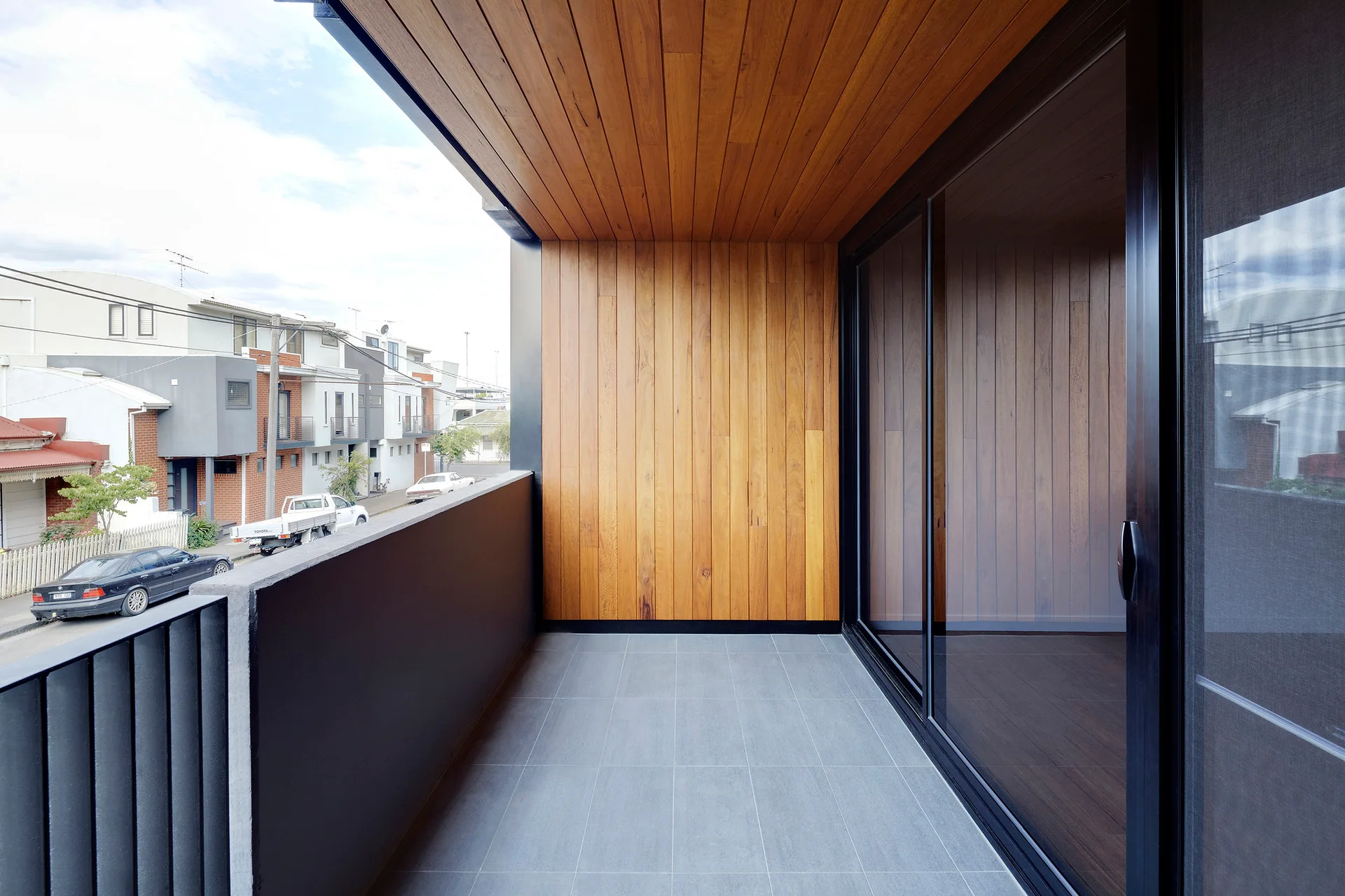 A modern balcony features warm vertical wood cladding on the wall and ceiling, grey tiled flooring, and a black-framed glass sliding door.