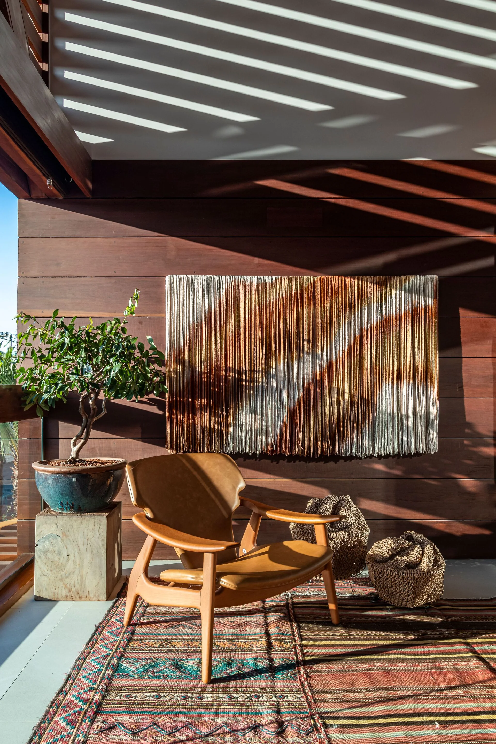 A mid-century leather lounge chair sits on a patterned rug on a wood-walled patio, beneath a large yarn wall hanging and slatted roof shadows.
