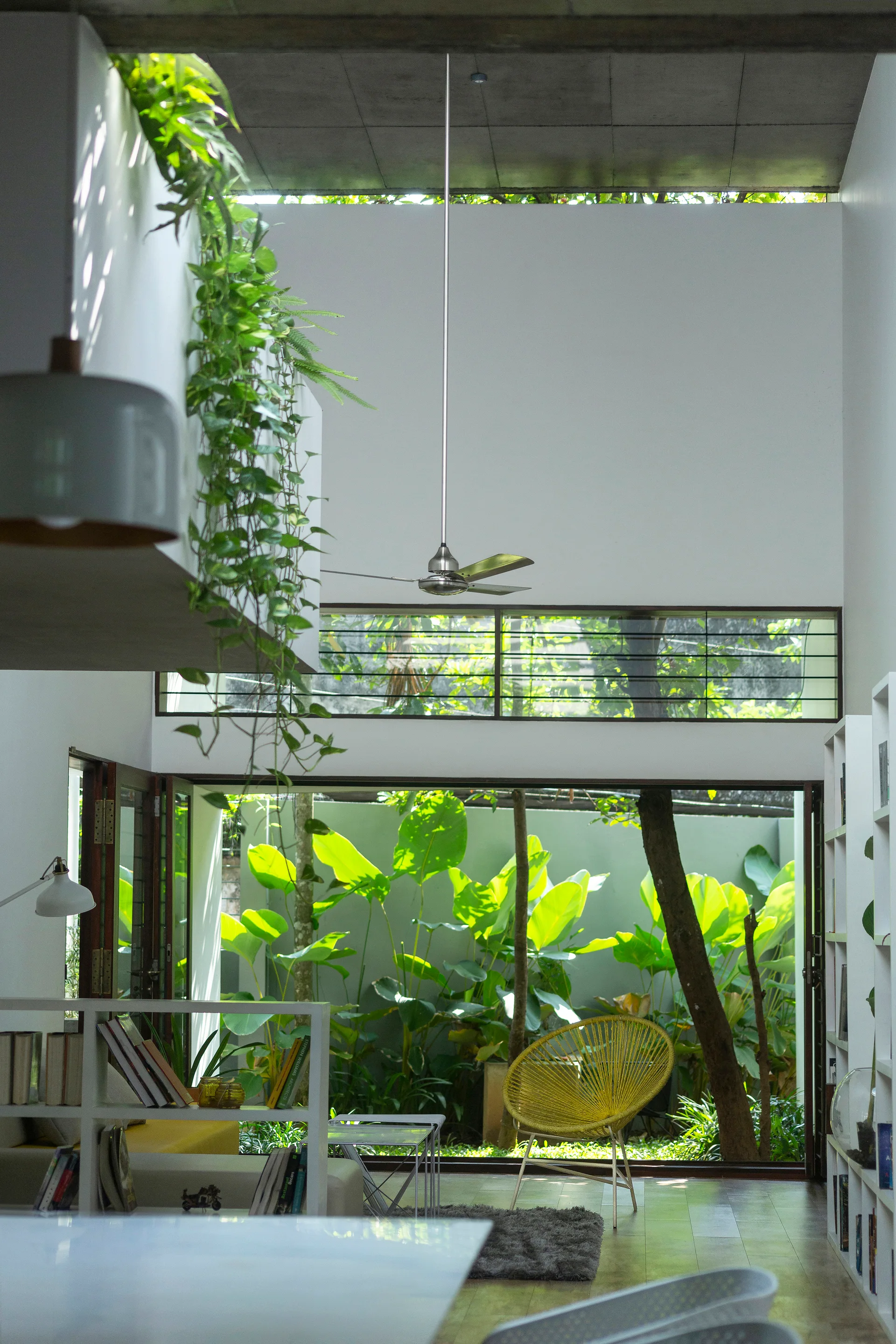 A high-ceilinged modern living room features a yellow Acapulco chair facing a large glass doorway that opens to a lush tropical courtyard garden.