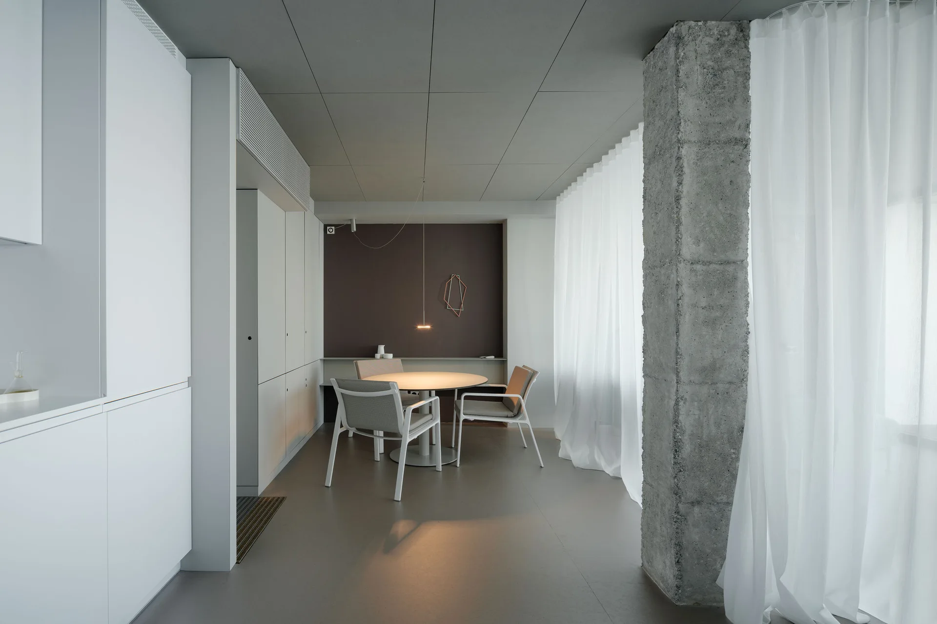 A minimalist dining area featuring a round table, white chairs, a dark accent wall, a rough concrete pillar, and flowing sheer white curtains.