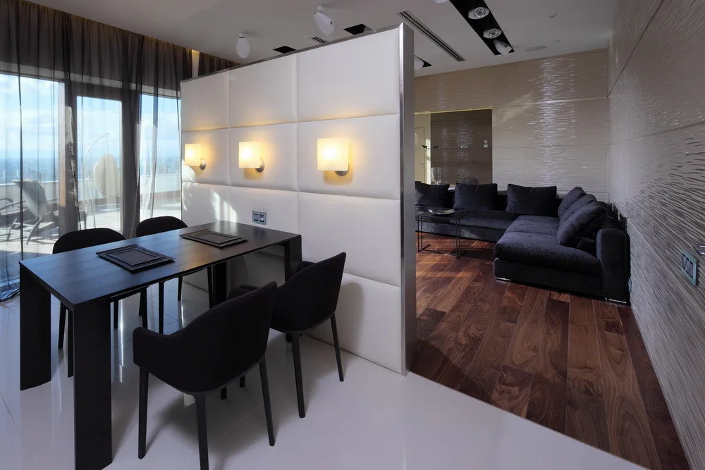A dark wood dining table and chairs sit next to a white upholstered partition wall with glowing sconces, separating the dining space from a living area.