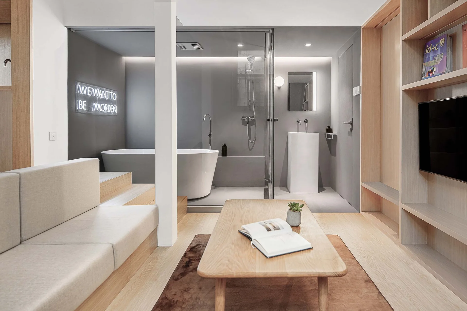 A minimalist living room with wood flooring and built-in shelving next to a glass-enclosed bathroom featuring a freestanding tub and pedestal sink.