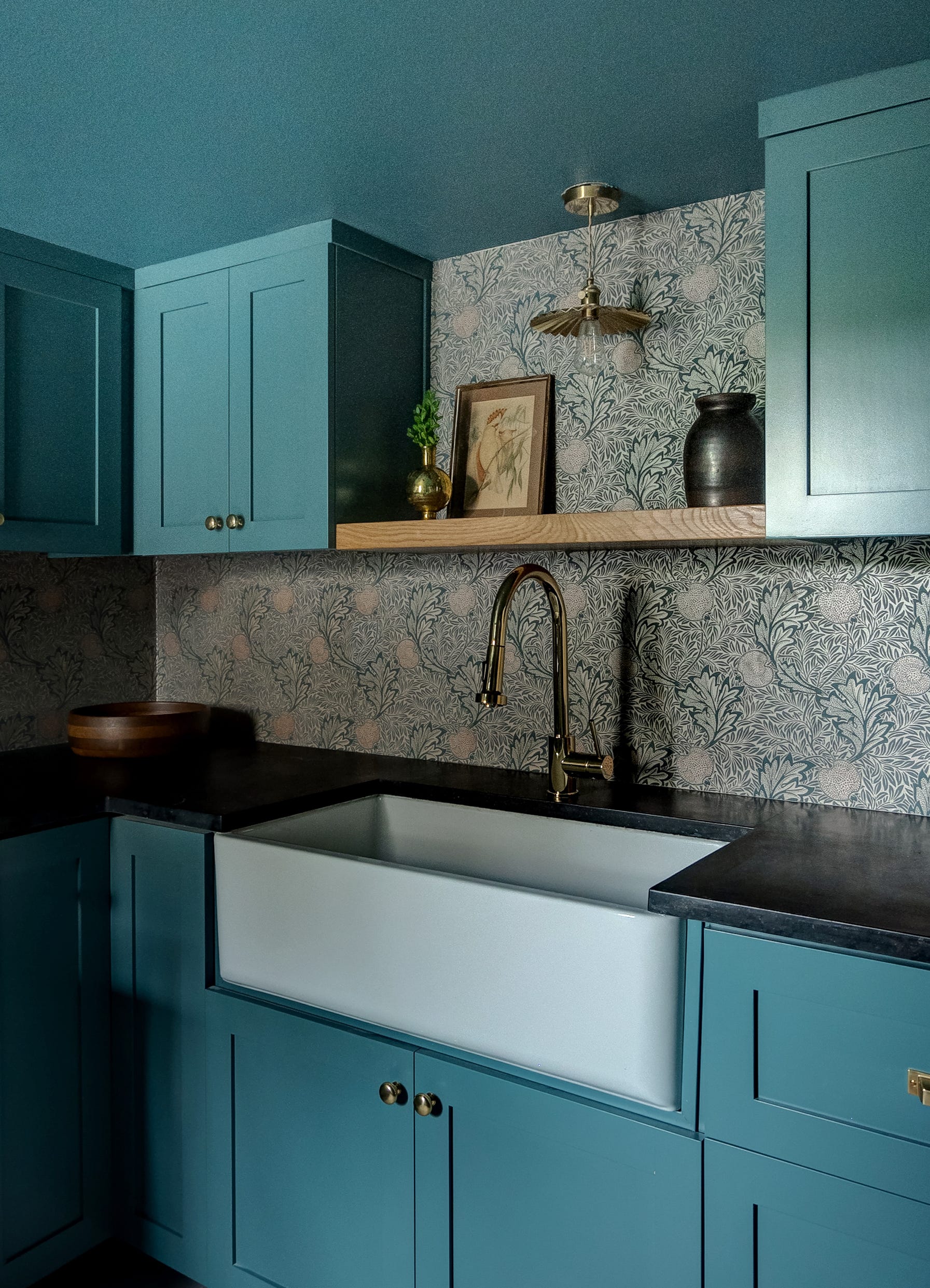 A white farmhouse sink with a brass faucet is set against dark countertops, teal shaker cabinets, and botanical print wallpaper.