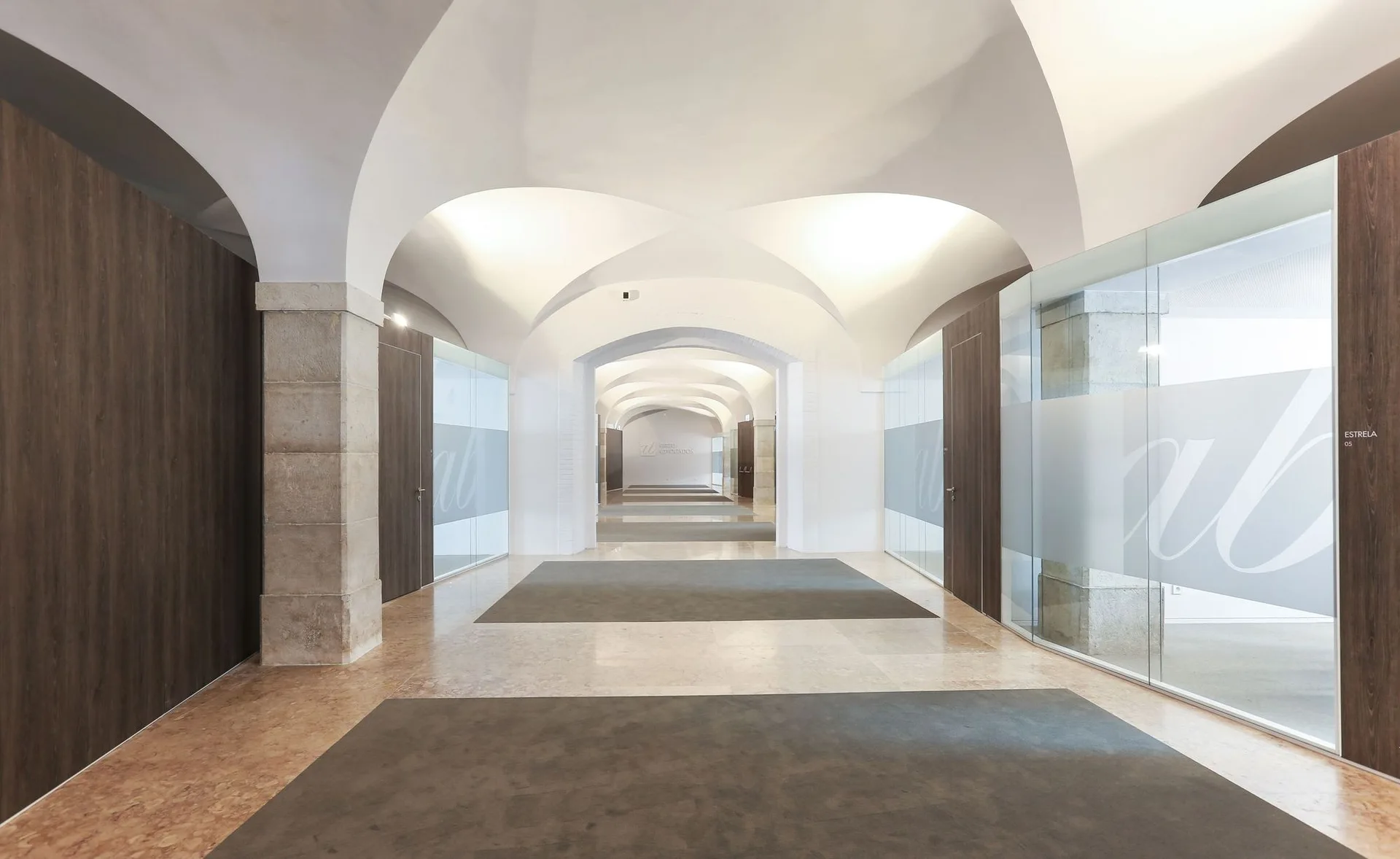 A symmetrical office hallway features a white vaulted ceiling, stone support pillars, glass partition walls, and dark wood doors.