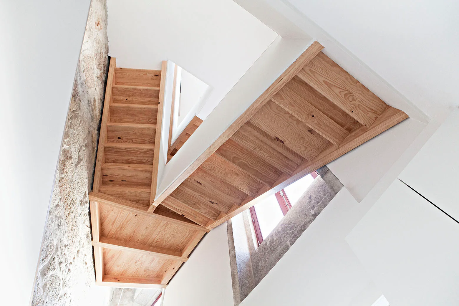 Looking up a multi-level wooden staircase built between a smooth white plaster wall and a rough, textured natural stone wall.