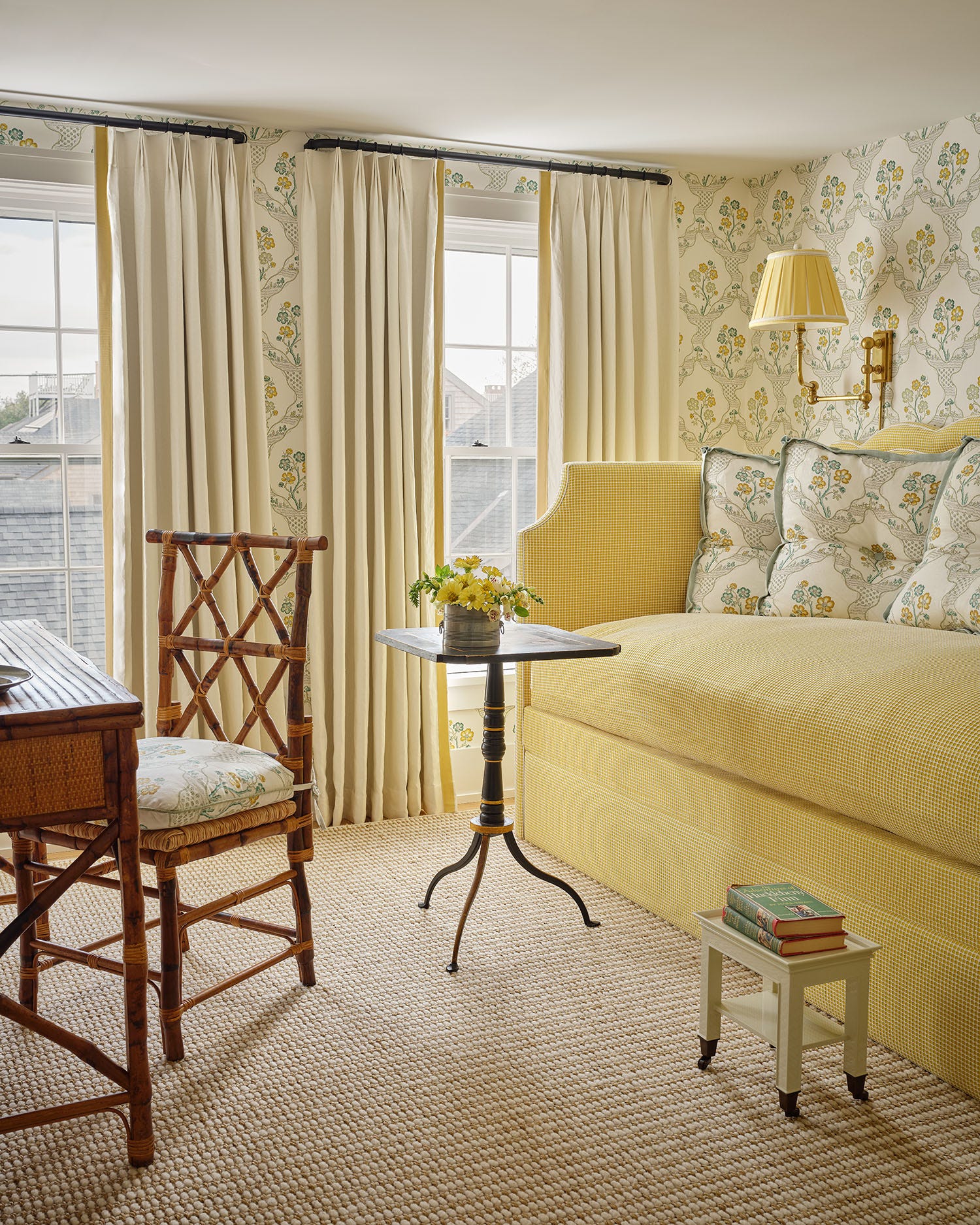 A cottage bedroom features a yellow gingham daybed, floral wallpaper, and a bamboo desk chair on a woven carpet.