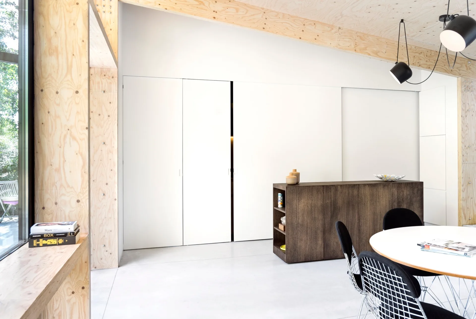 A minimalist dining room features a white round table with wire chairs, a dark wood console, and large white sliding doors under a plywood ceiling.