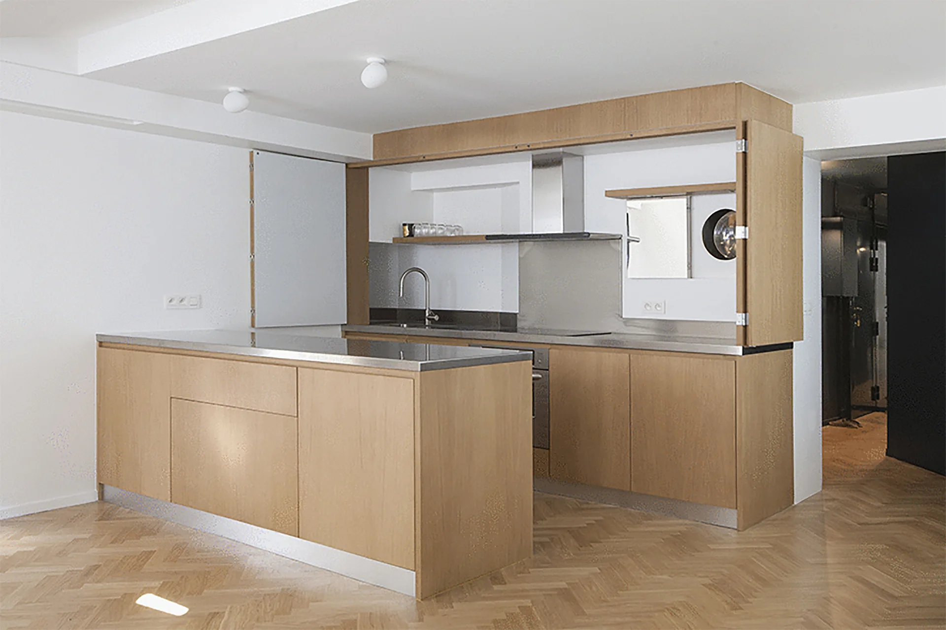 A minimalist kitchen features a light wood island, stainless steel countertops, and a herringbone pattern wood floor.