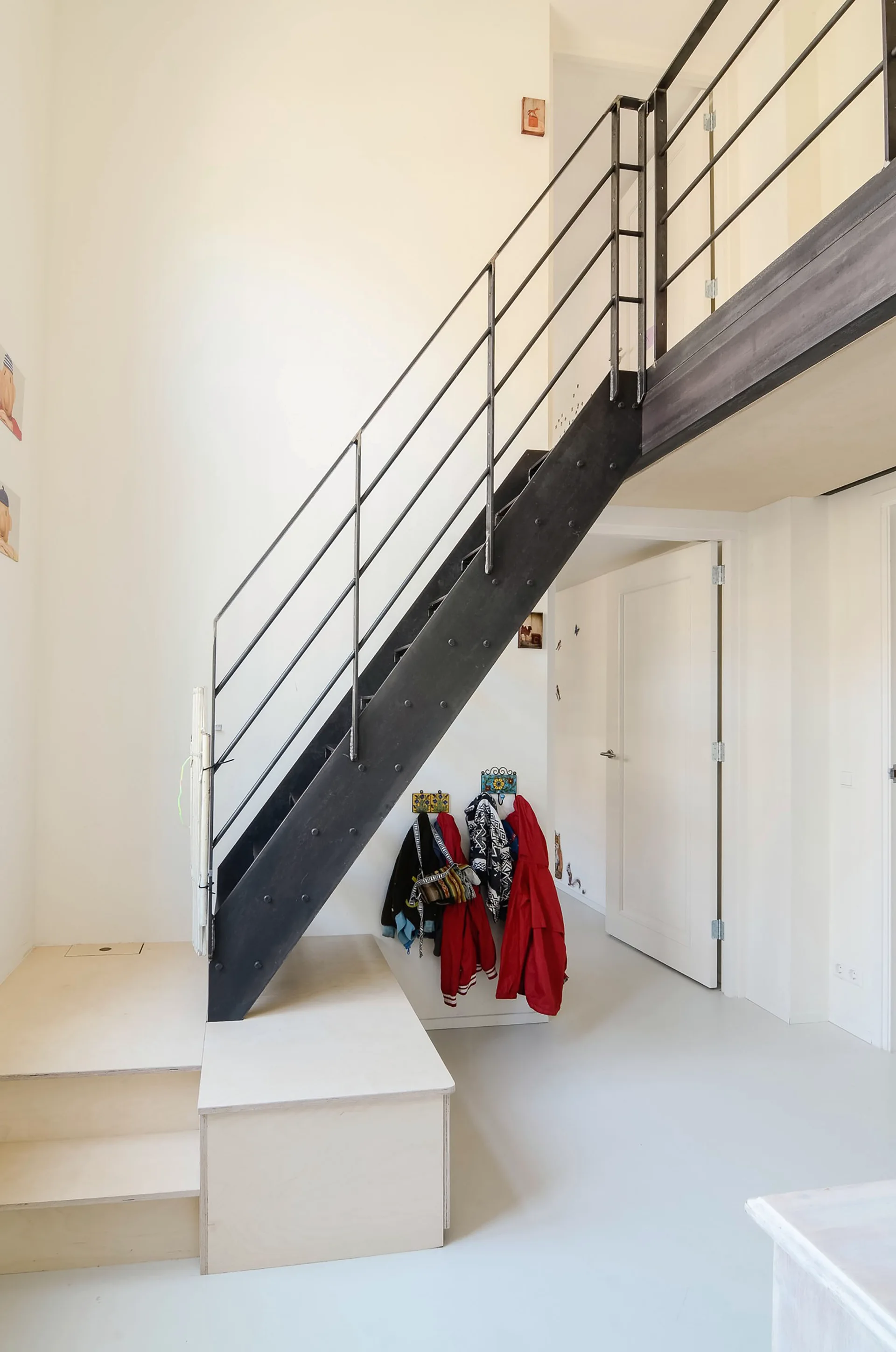 A black metal industrial staircase with thin railings stands on a light plywood platform base in a minimalist white hallway with coats hanging underneath.