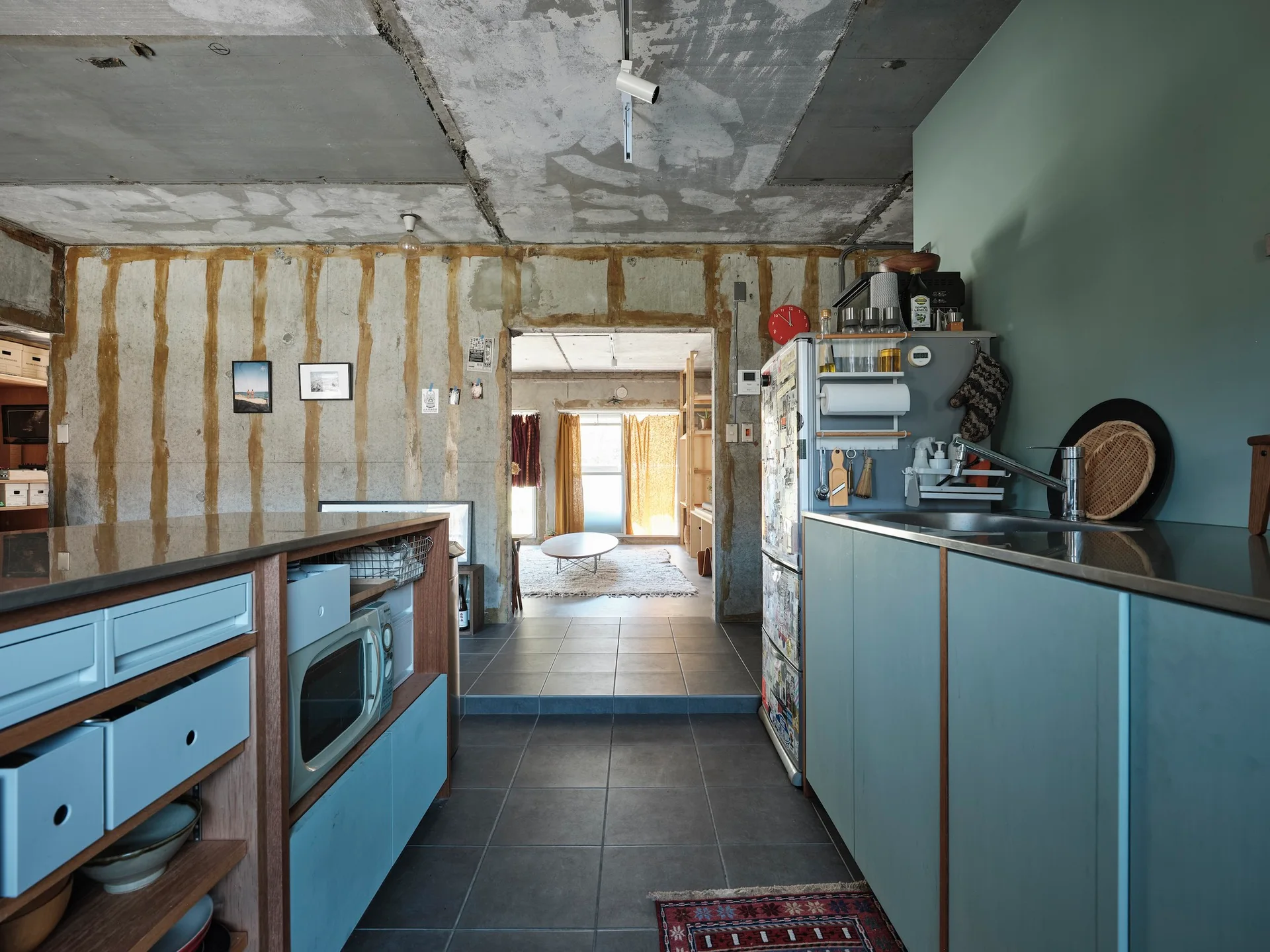 An industrial kitchen features matte blue cabinetry and a concrete island set against raw, unfinished concrete walls and grey tiled floors.