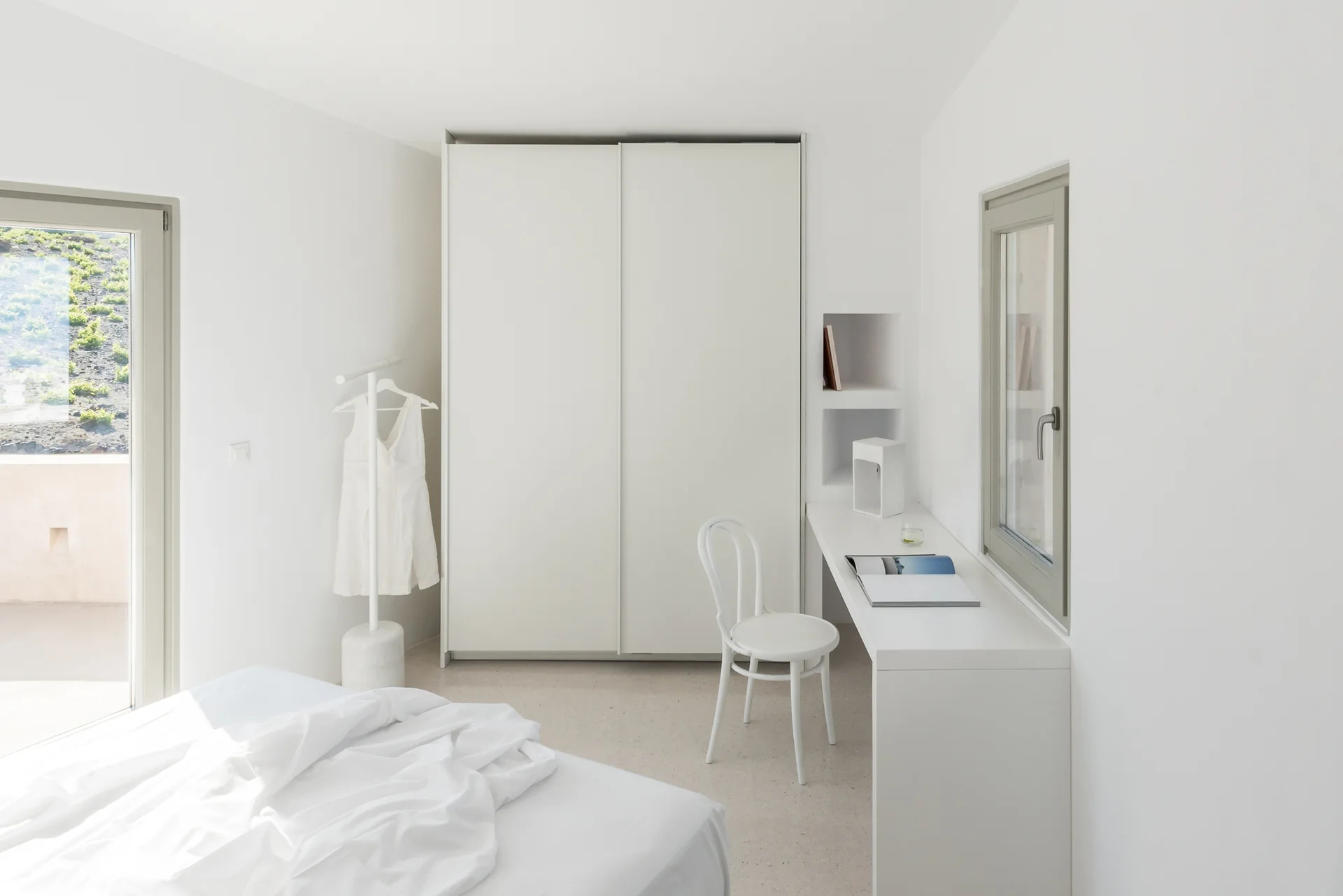 A minimalist white bedroom featuring a built-in desk under a window, a white bistro chair, a tall sliding wardrobe, and a bed with rumpled linens.