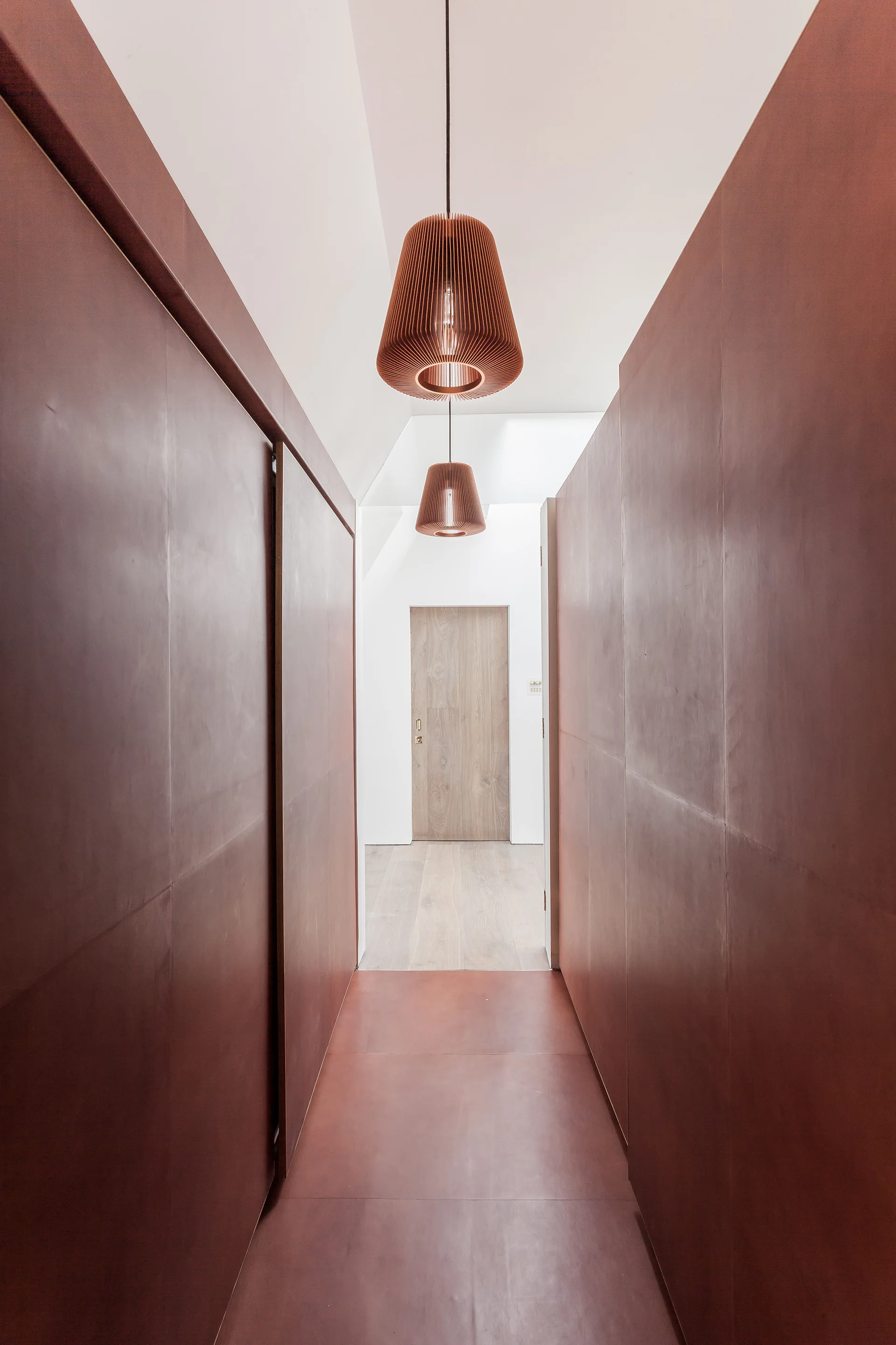 A narrow hallway with matte burgundy paneled walls and floors features two copper-colored pendant lights leading toward a light wood door.