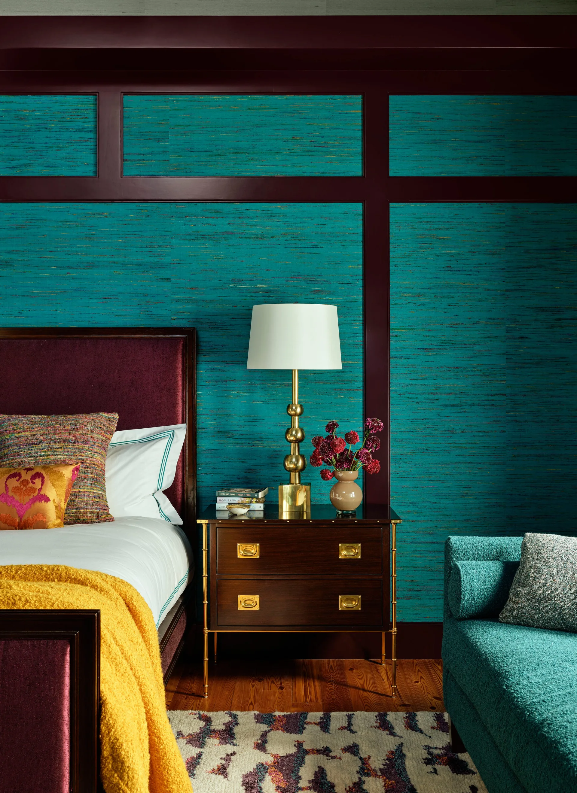 A bedroom features a bed with a burgundy headboard and a dark wood nightstand against textured teal wallpaper with burgundy painted trim.