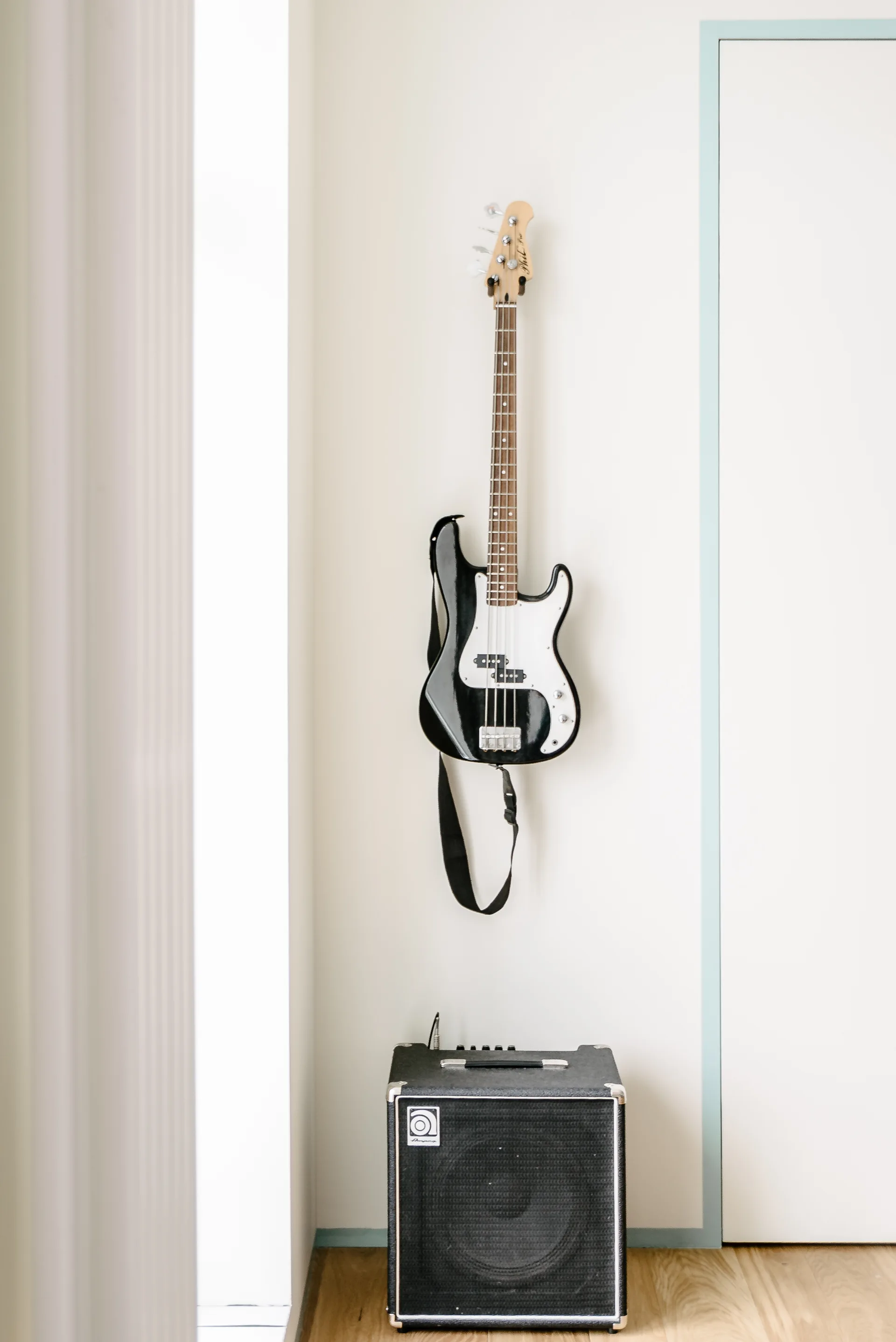 A black bass guitar hangs on a white wall above a matching black amplifier on a light wood floor.