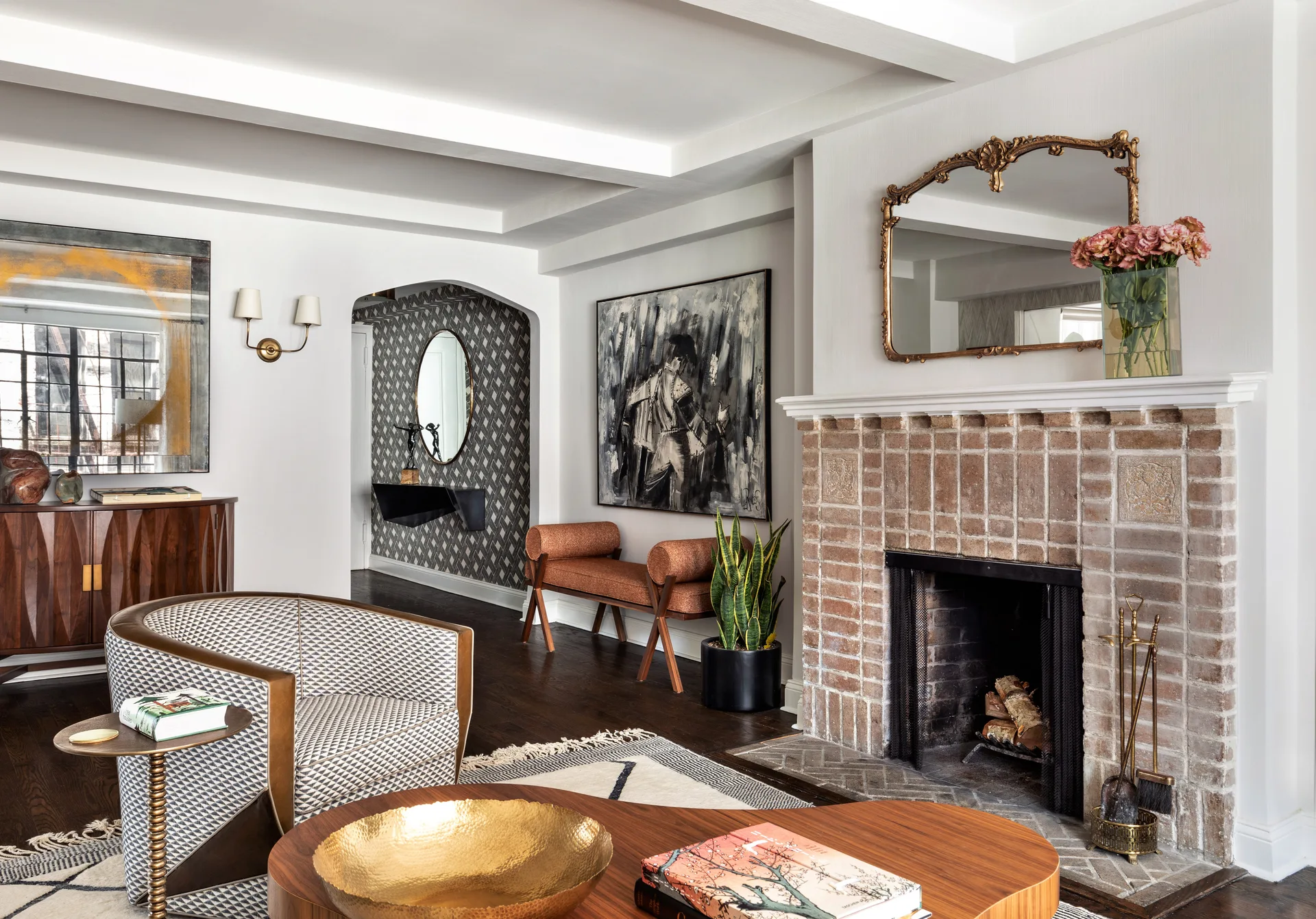A living room featuring a brick fireplace with an ornate gilded mirror above it, a patterned armchair, and an arched hallway with geometric wallpaper.