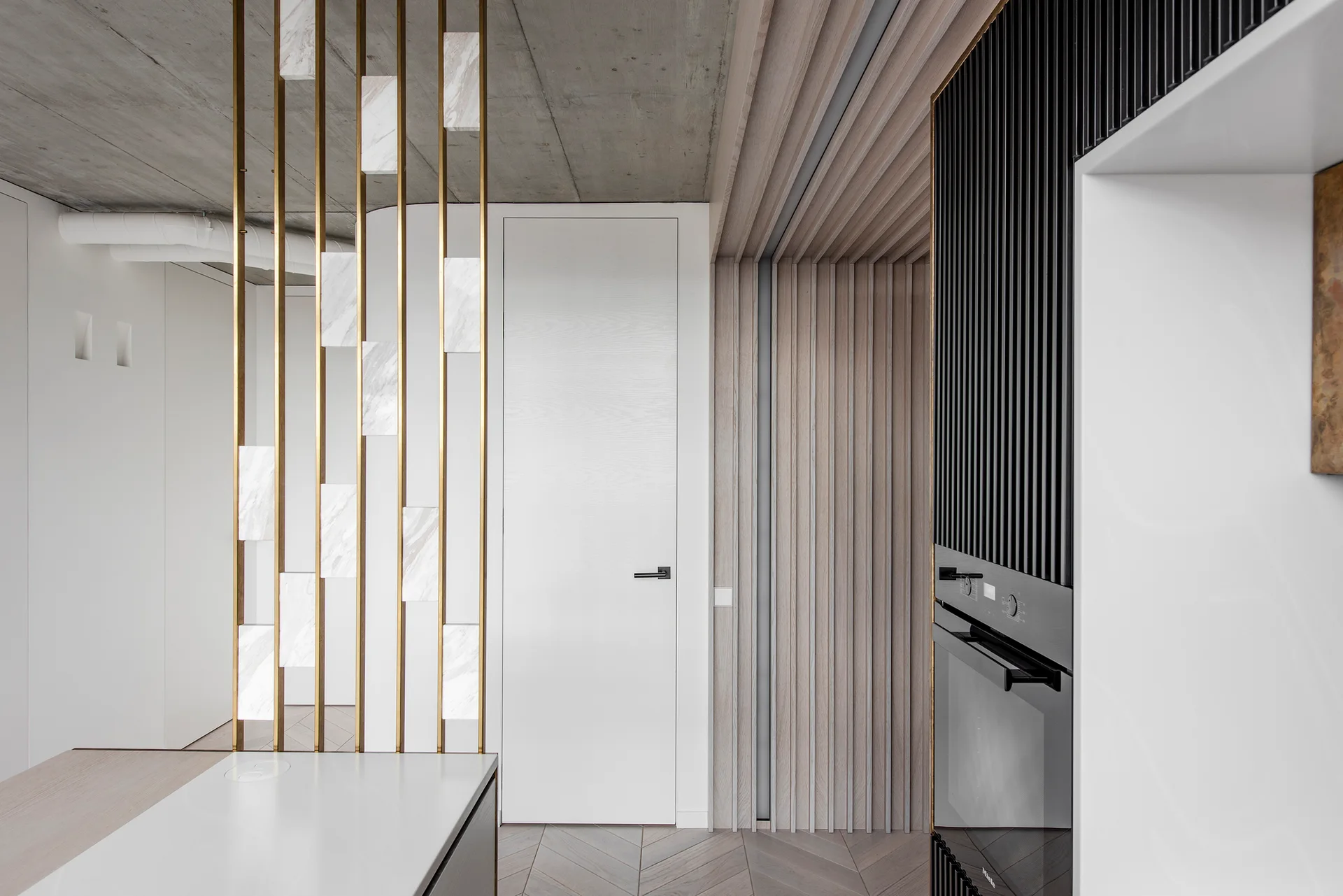 A modern kitchen interior featuring a custom brass and marble room divider next to chevron floors, vertical wood slats, and a white door.