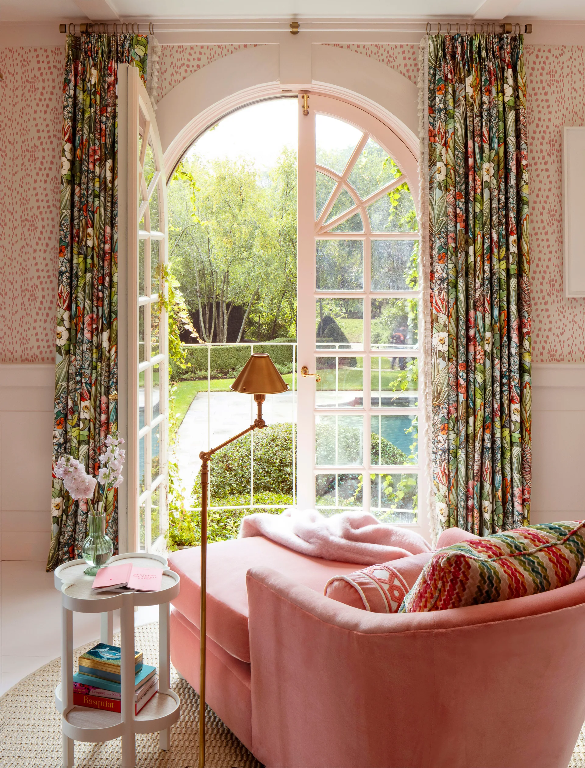 A pink chaise lounge sits next to a white tiered side table in front of an open arched French door with botanical-print curtains.