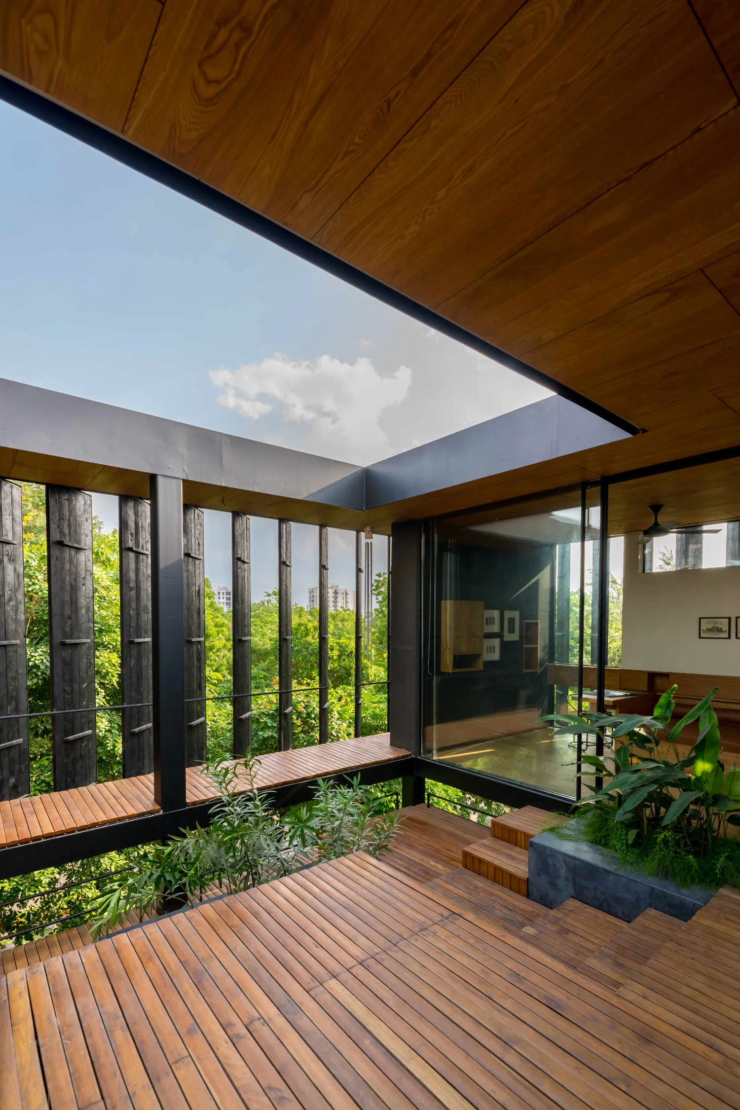 A contemporary wooden terrace deck features a large rectangular roof cutout, vertical black wood screens, a stone planter, and glass walls looking inside.