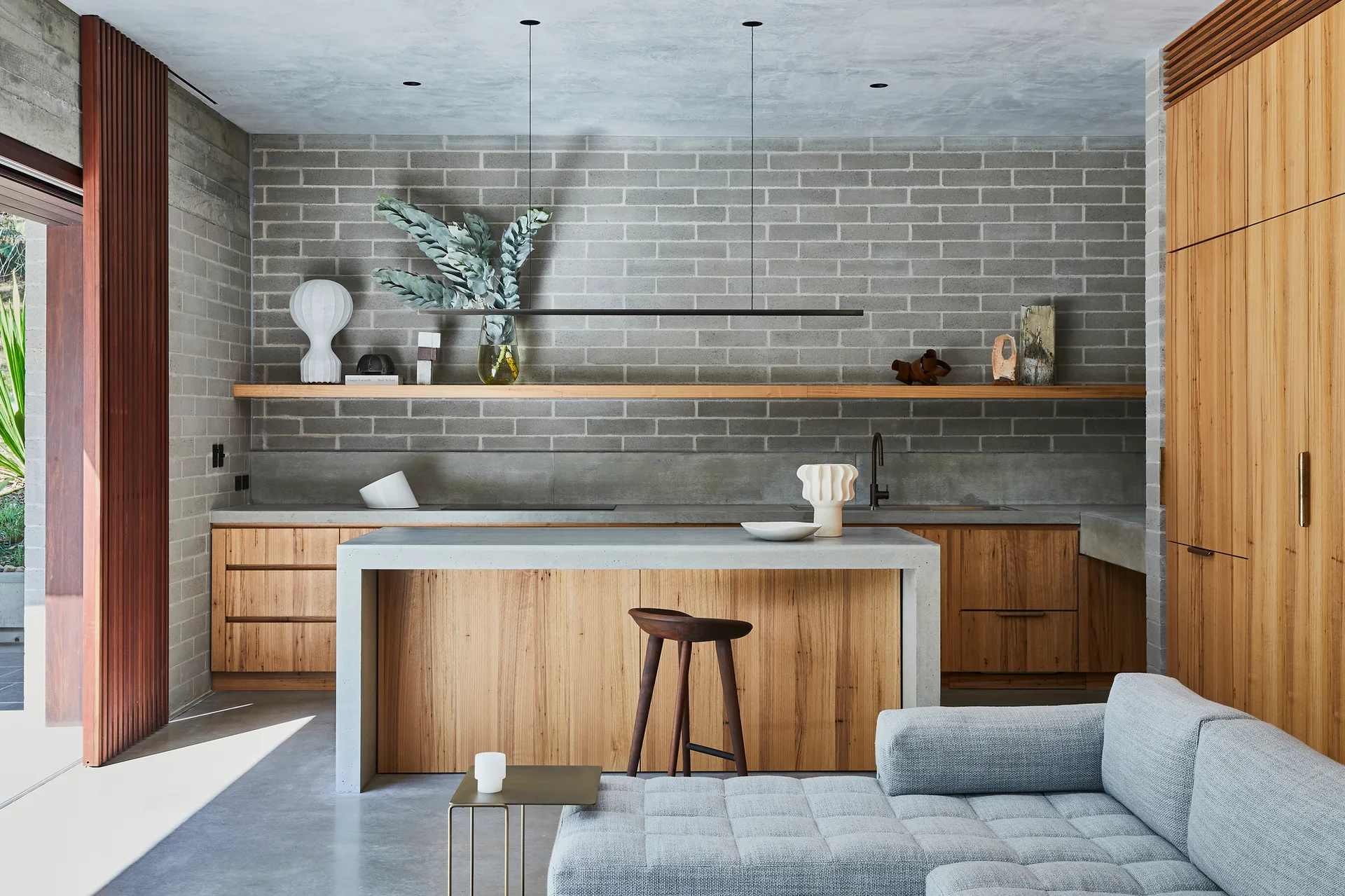 A modern kitchen features a concrete-wrapped wood island, flat-front wood cabinets, and an exposed grey brick accent wall under a long wooden shelf.