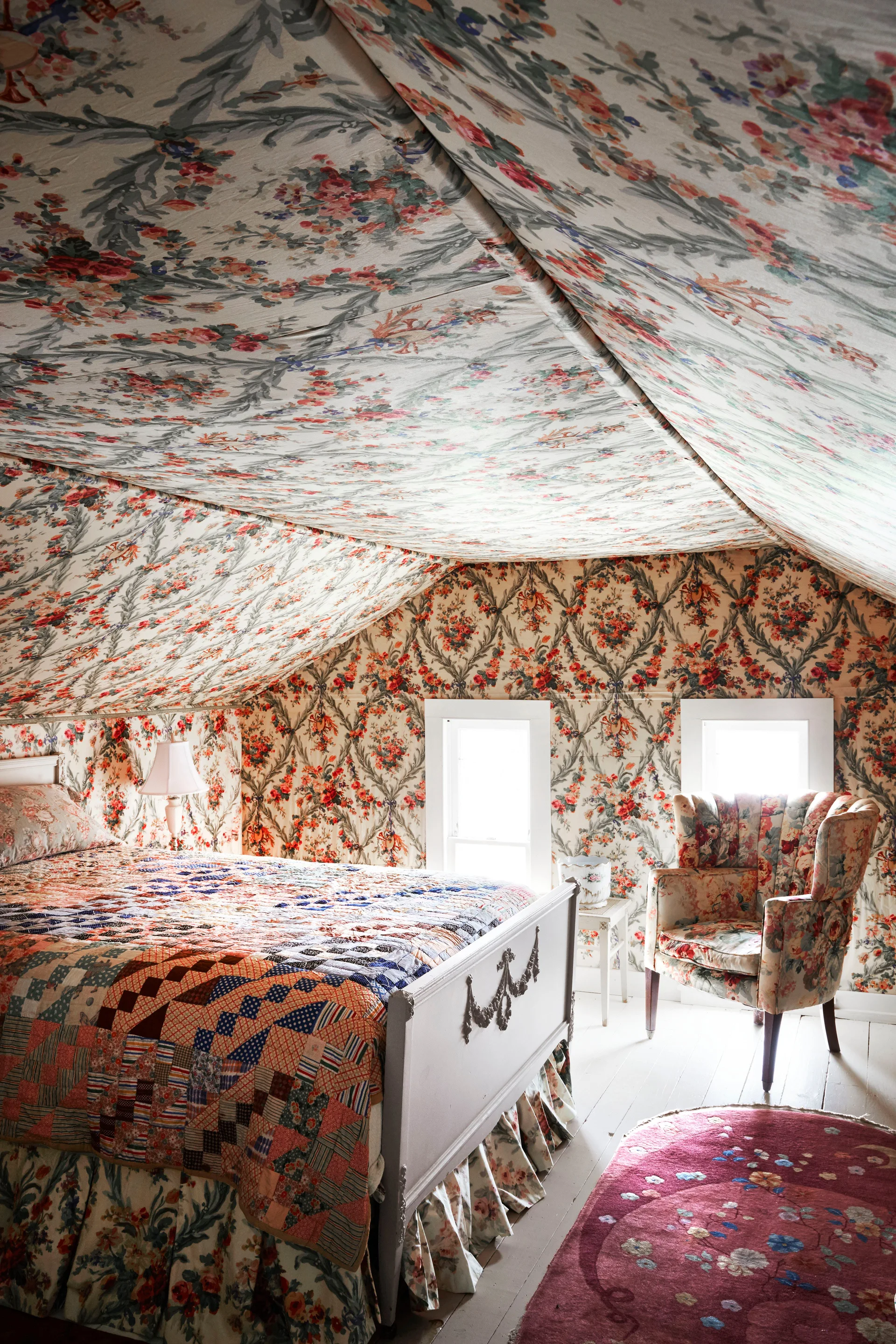 An attic bedroom features matching floral wallpaper on the walls and slanted ceiling, a wooden bed with a patchwork quilt, and a floral armchair.