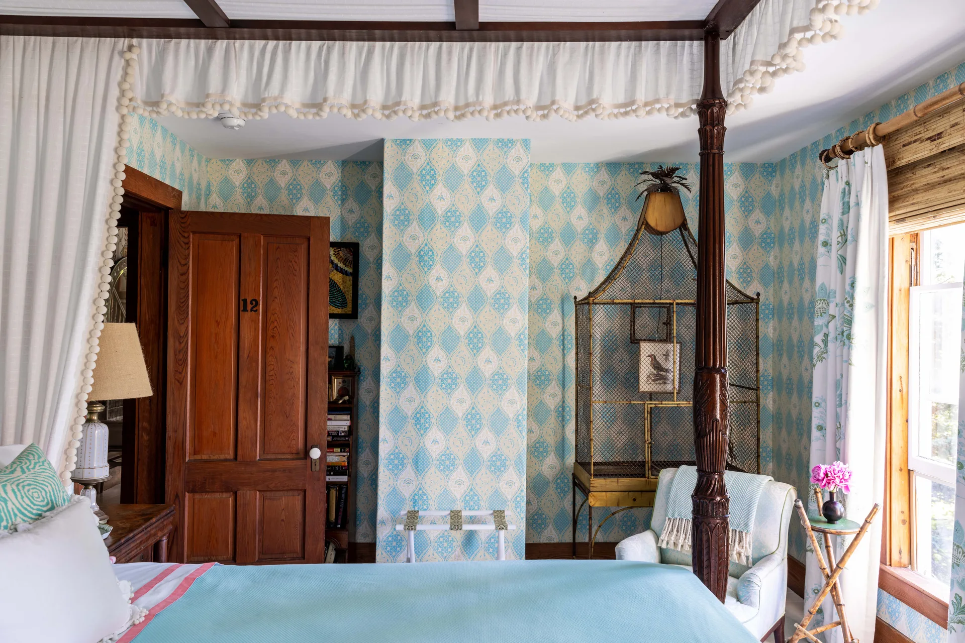 A bedroom features blue and cream lattice wallpaper, a dark wood canopy bed with pom-pom trim, and a vintage decorative bird cage.