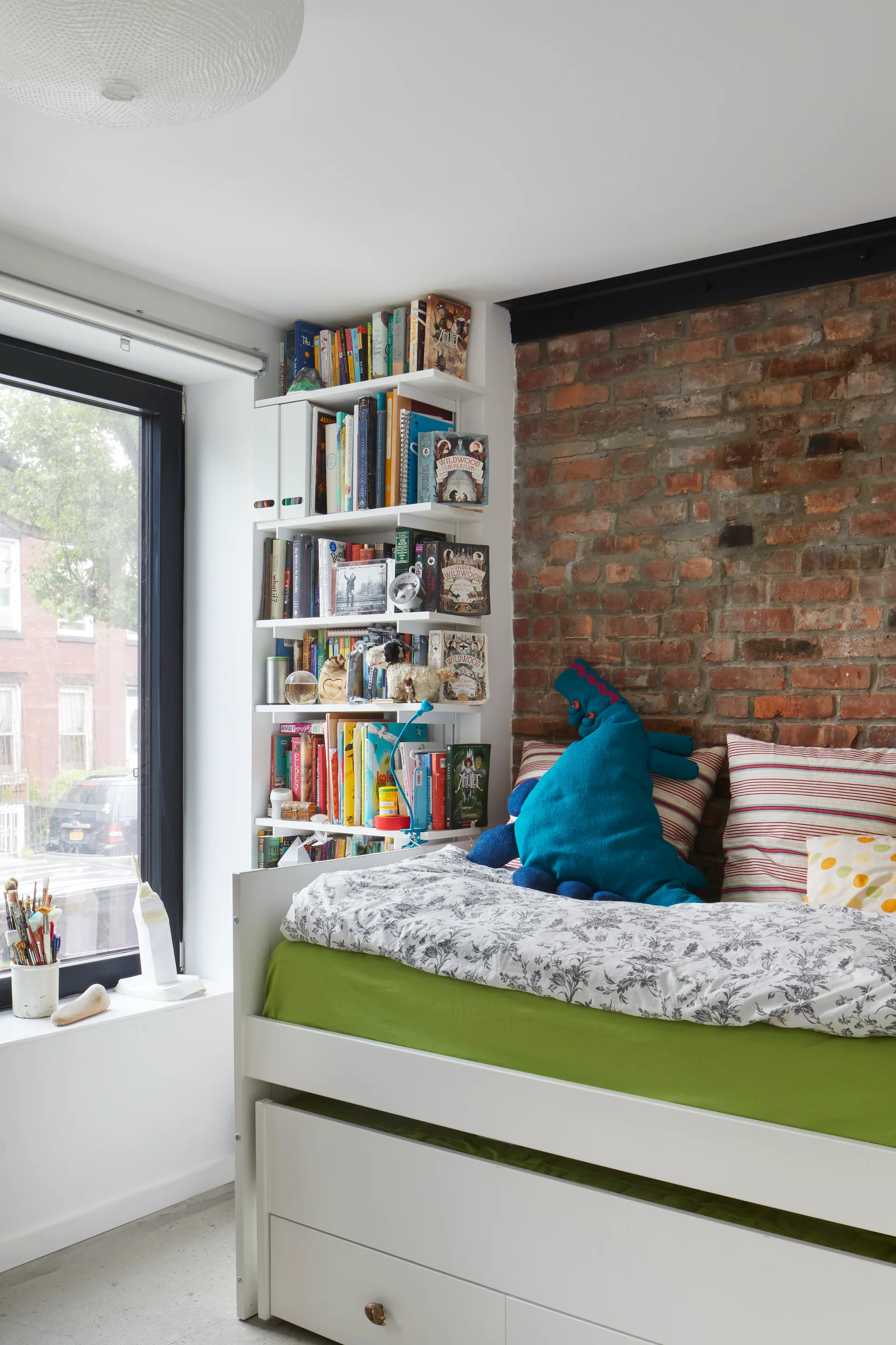 A child's white trundle bed with green sheets sits next to an exposed red brick wall and a tall white corner bookshelf packed with books.