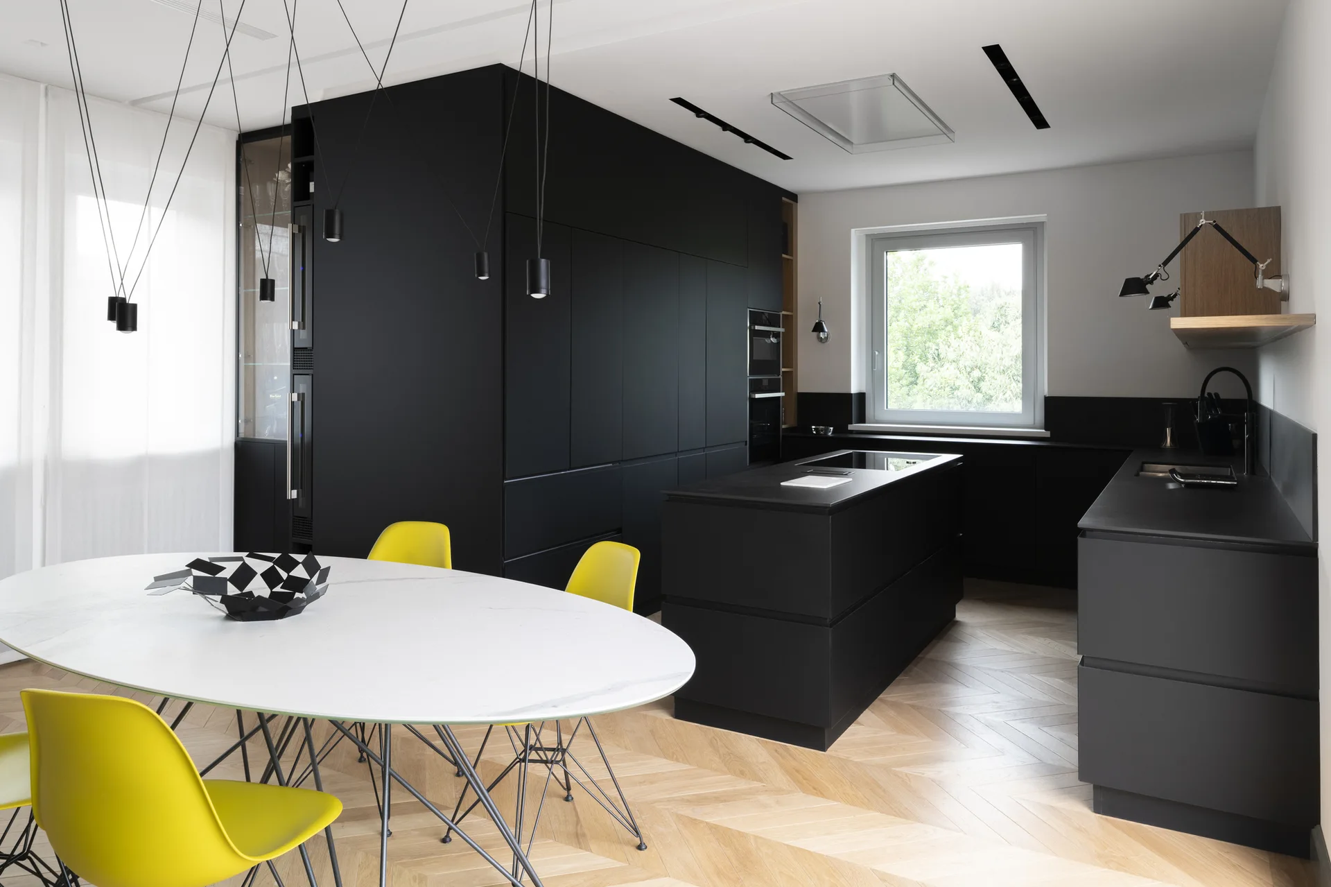 A modern kitchen featuring matte black cabinetry, a central island, and a white dining table with yellow chairs on herringbone wood floors.