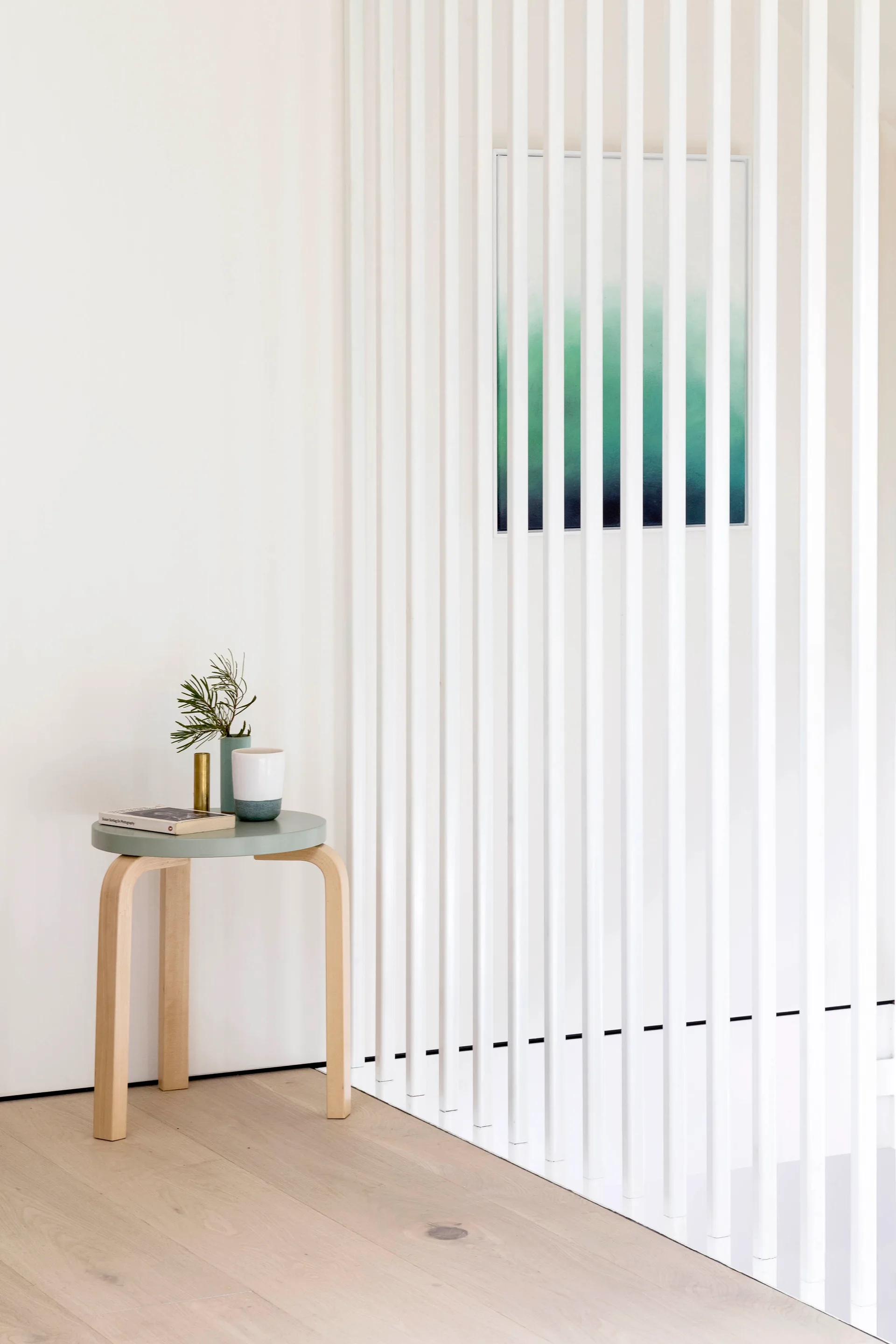 A round sage green side table with decorative vases stands next to a white vertical slatted stair screen on light hardwood floors.