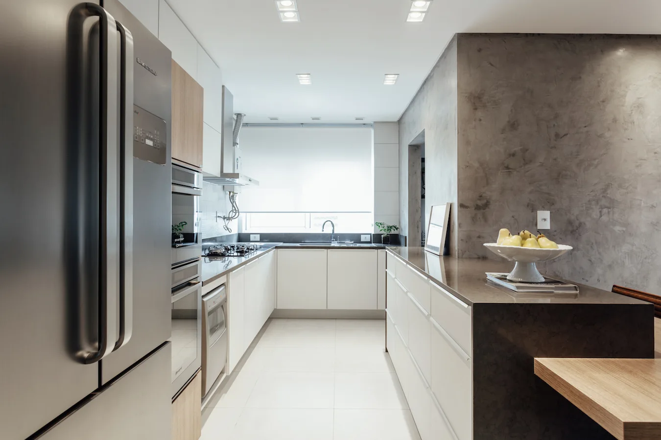A modern galley kitchen with white and wood cabinets, a stainless steel refrigerator, white tile floors, and a textured grey concrete wall.