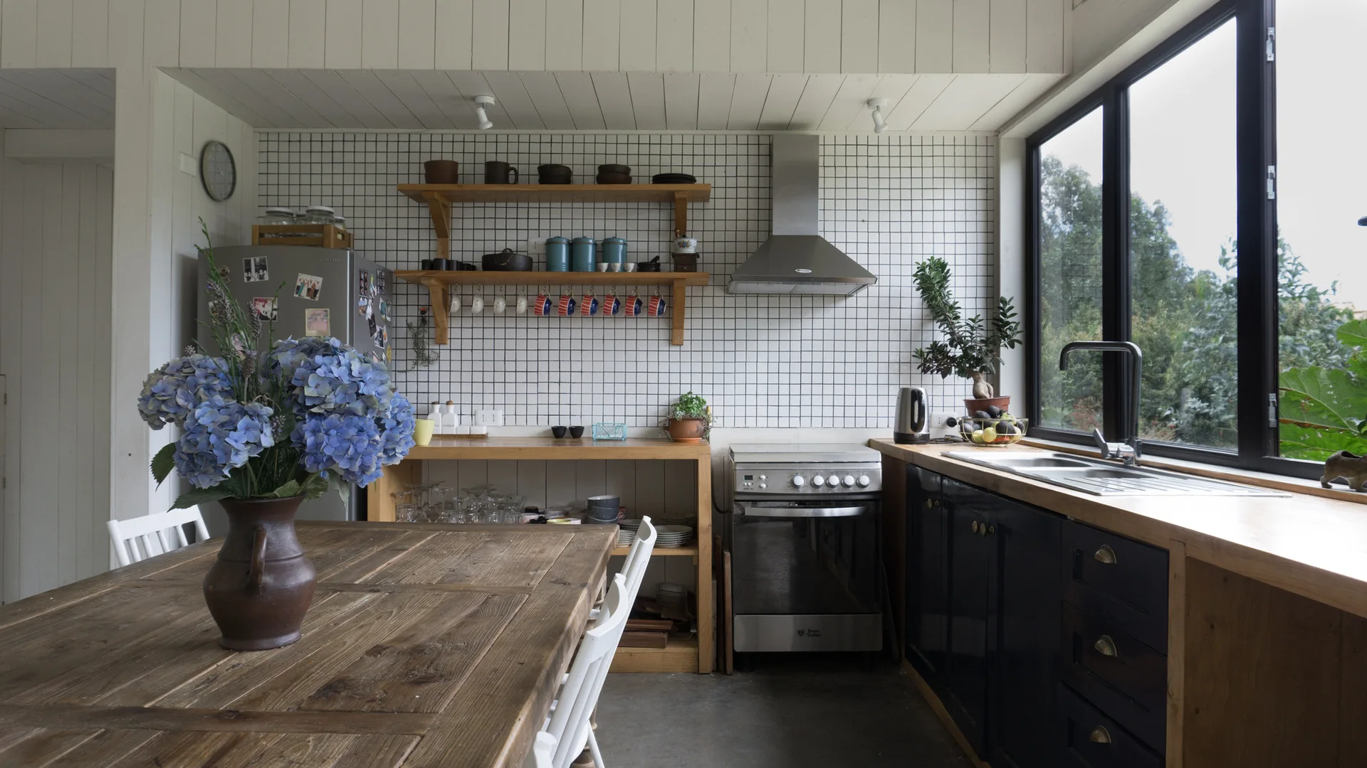A rustic kitchen featuring a white grid tile wall, dark blue base cabinets with wood countertops, and a wooden dining table with blue hydrangeas.