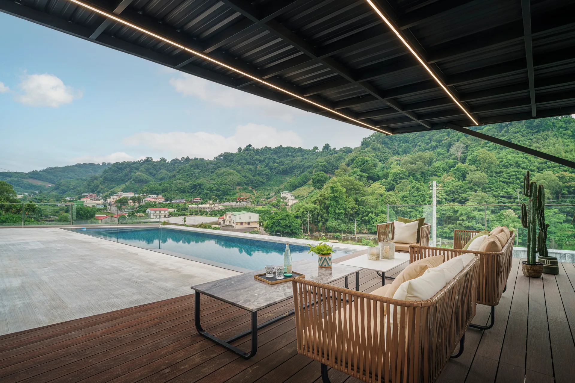 A covered wooden patio features woven outdoor lounge furniture, a stone coffee table, and an infinity pool looking out at green hills.