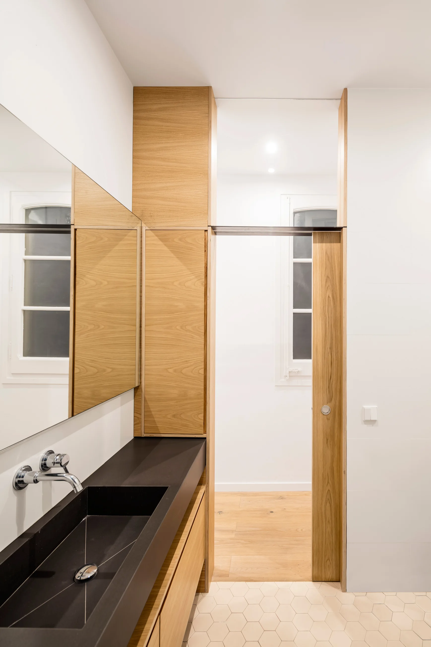 A modern minimalist bathroom featuring a black stone vanity with an integrated basin, custom light oak cabinetry, and beige hexagonal floor tiles.