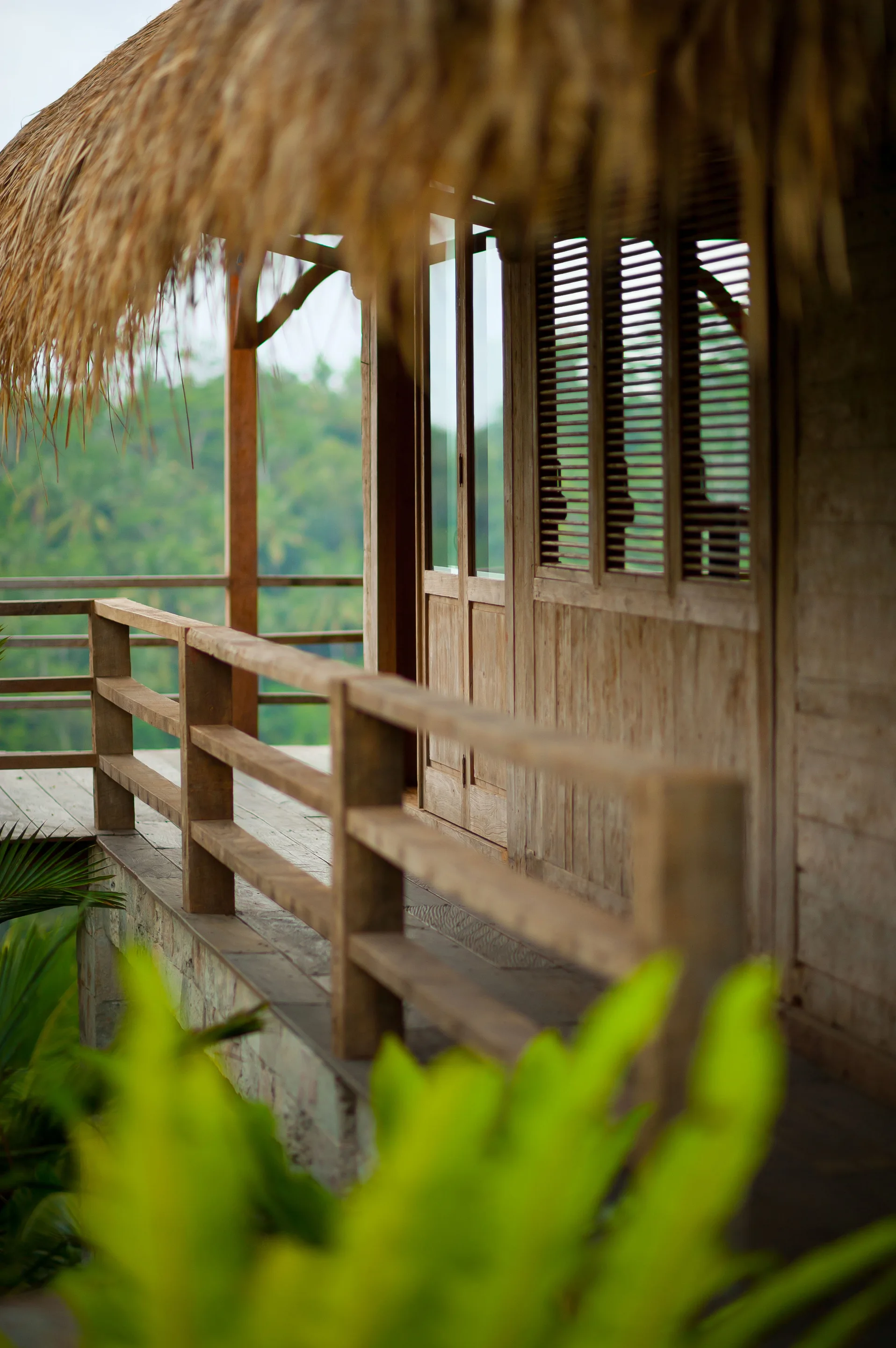 A rustic wooden veranda features a weathered timber railing, matching wood plank walls with slatted shutters, and a textured thatched roof.