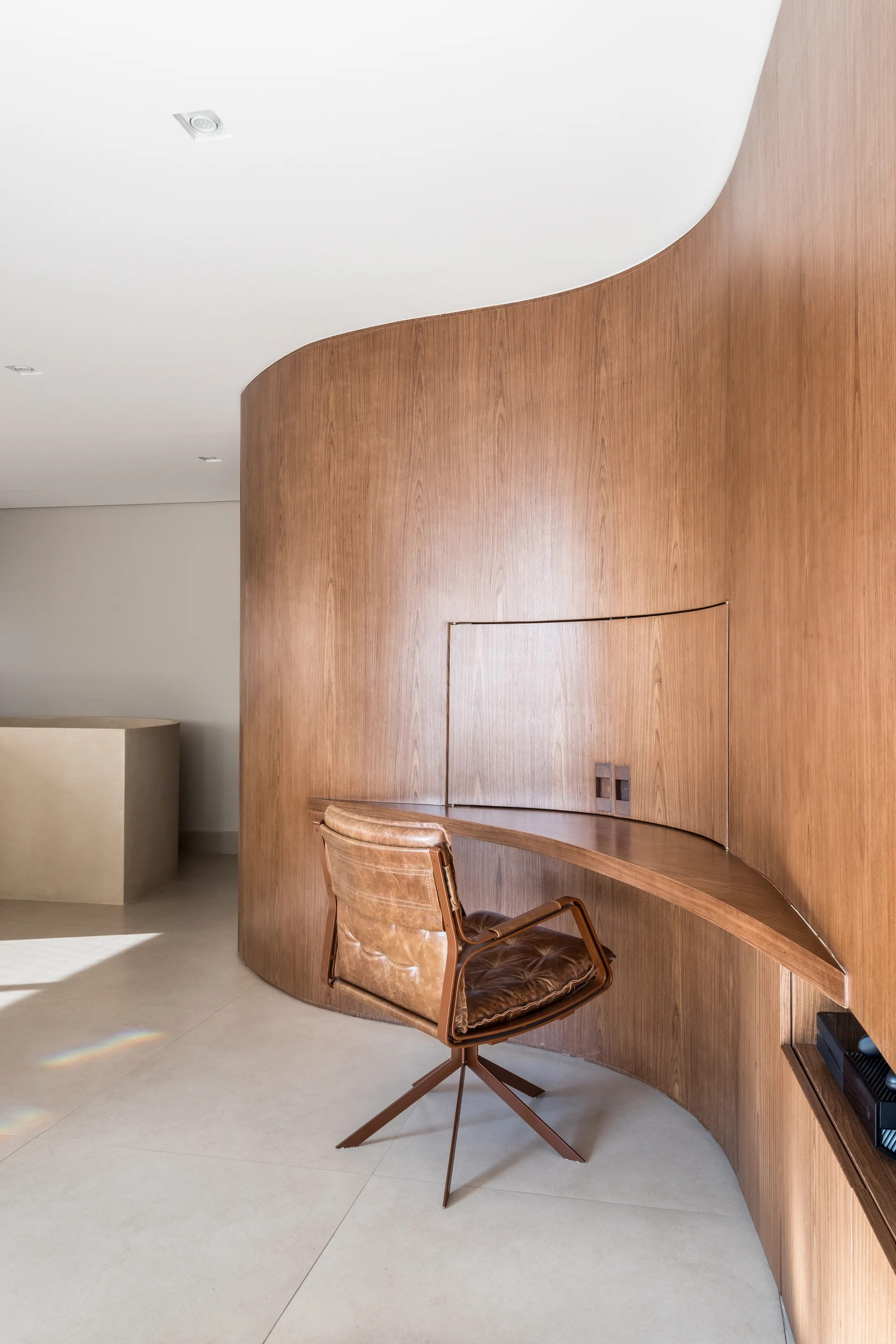 A contemporary home office features a curved wood-clad wall with an integrated floating desk and a brown leather swivel chair on light tile flooring.
