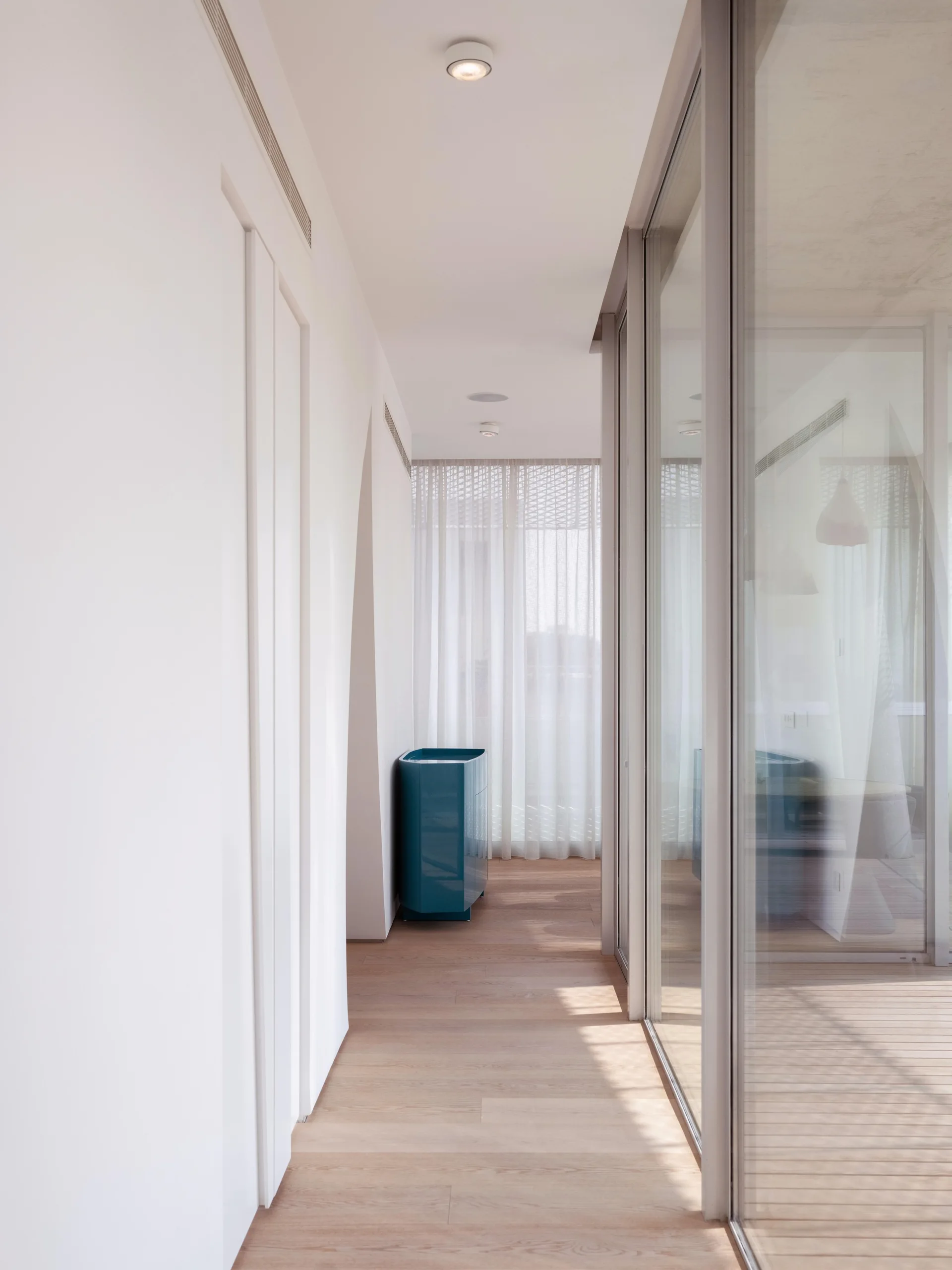 A bright minimalist hallway featuring light wood flooring, glass wall partitions, and a single glossy blue cabinet set against sheer white curtains.