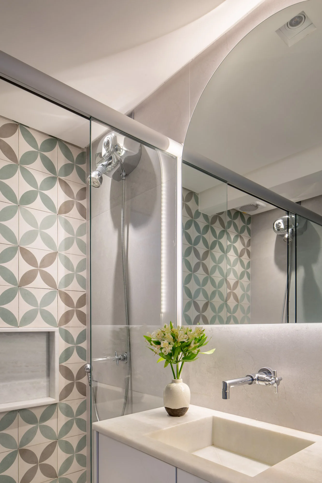 A bathroom vanity with an integrated sink and wall-mounted faucet features an arched mirror alongside a glass shower panel with sage and grey geometric tiles.