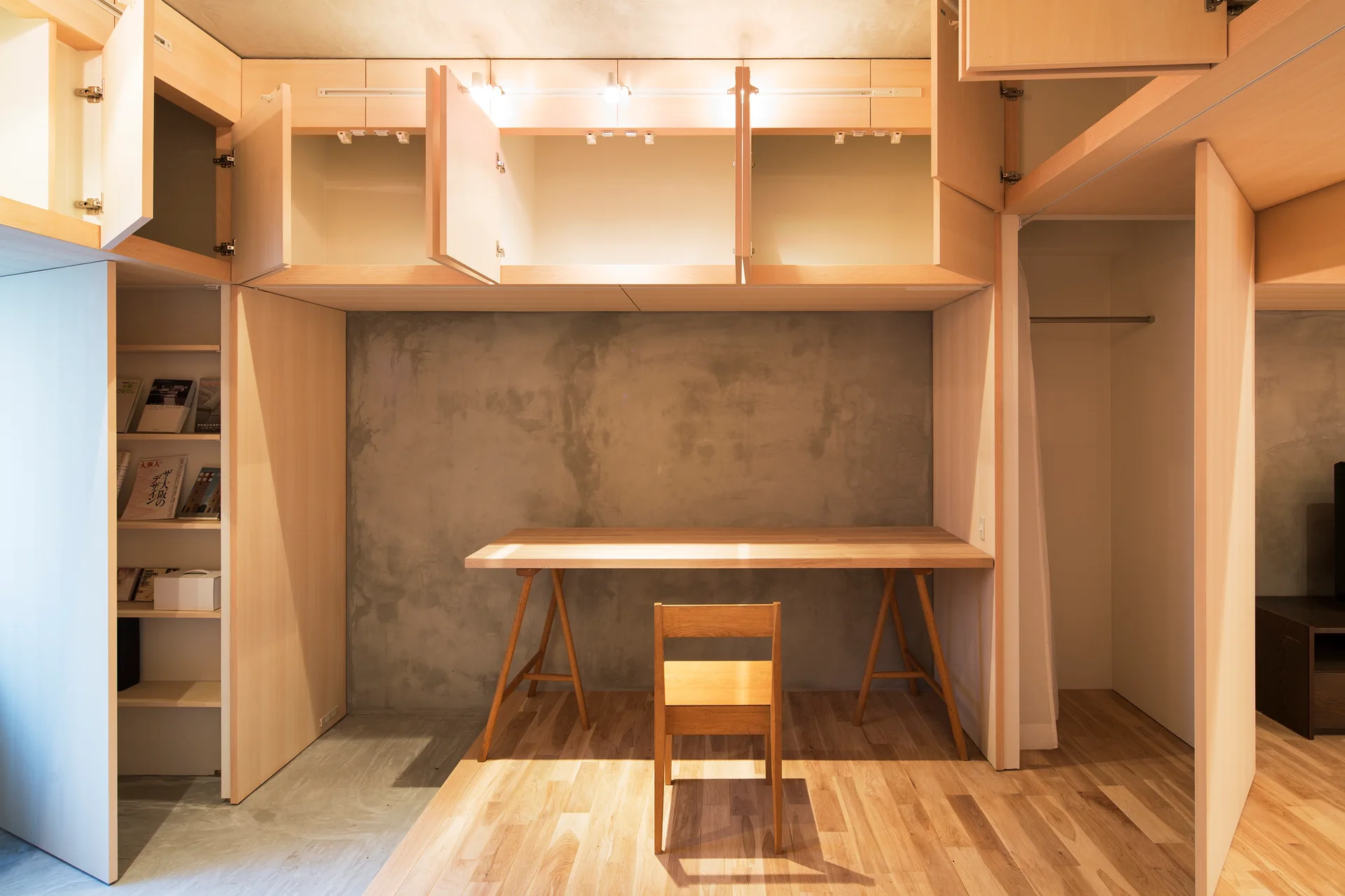 A minimalist home office features a light wood trestle desk and chair set against a raw grey concrete wall under built-in wooden cabinets.