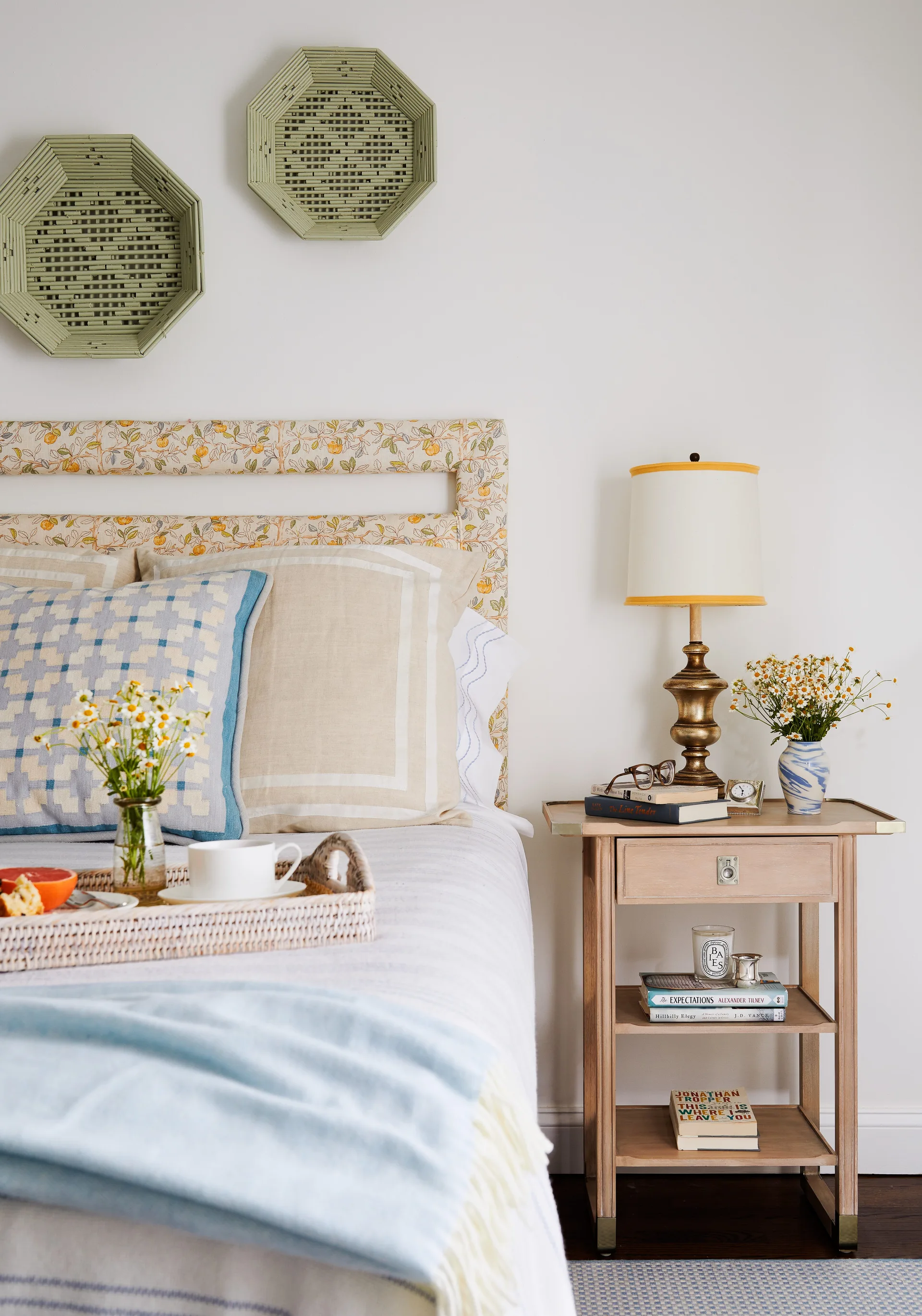 A cozy bedroom featuring a floral print upholstered headboard, a light wood nightstand with a brass lamp, and a wicker tray on the bed.