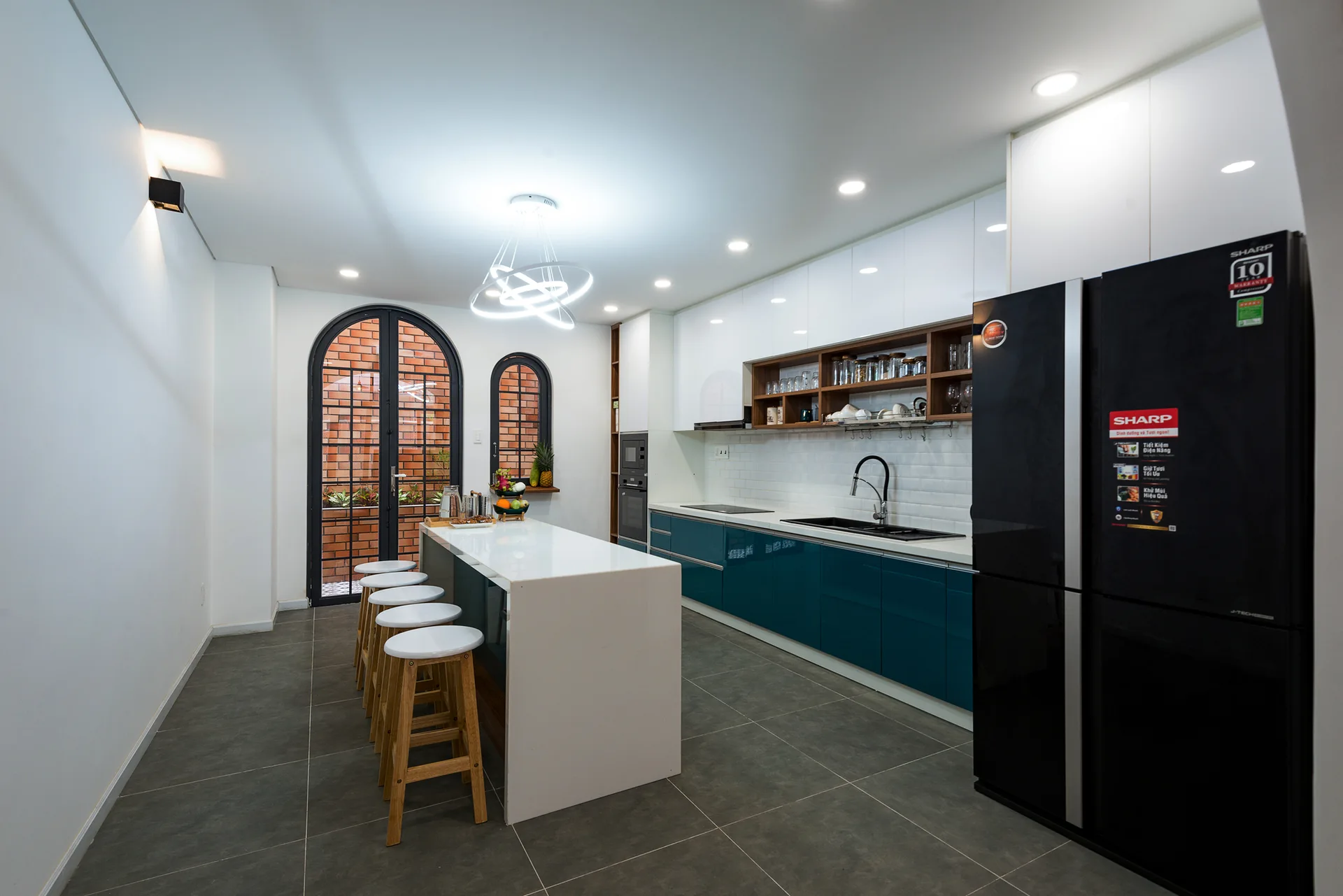 A modern kitchen features a white central island with wooden stools, glossy teal lower cabinets, and arched glass doors looking out to a brick wall.