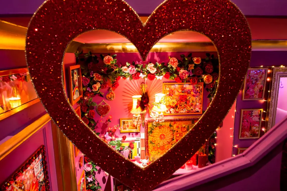 A bright fuchsia wall featuring gold-framed art and rose garlands is viewed through a large red glitter heart frame.