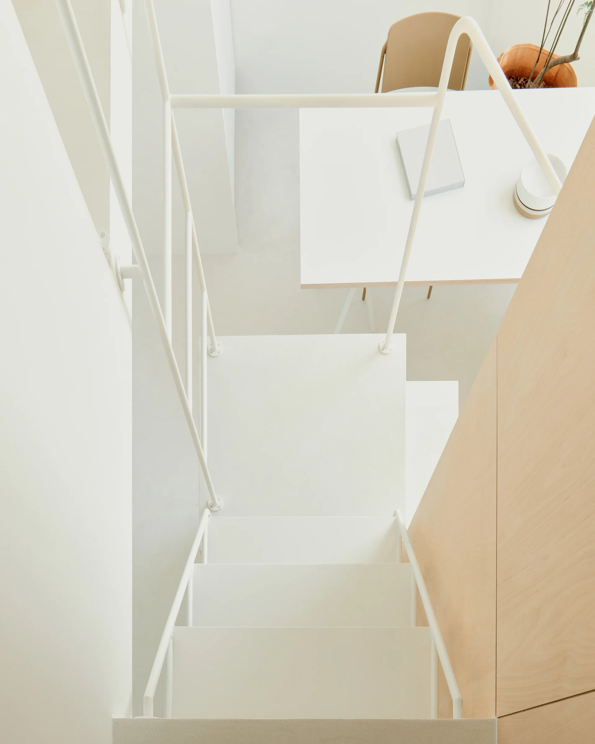 An overhead view down a white metal staircase looking onto a white desk, a light wood paneled wall, and a beige chair.
