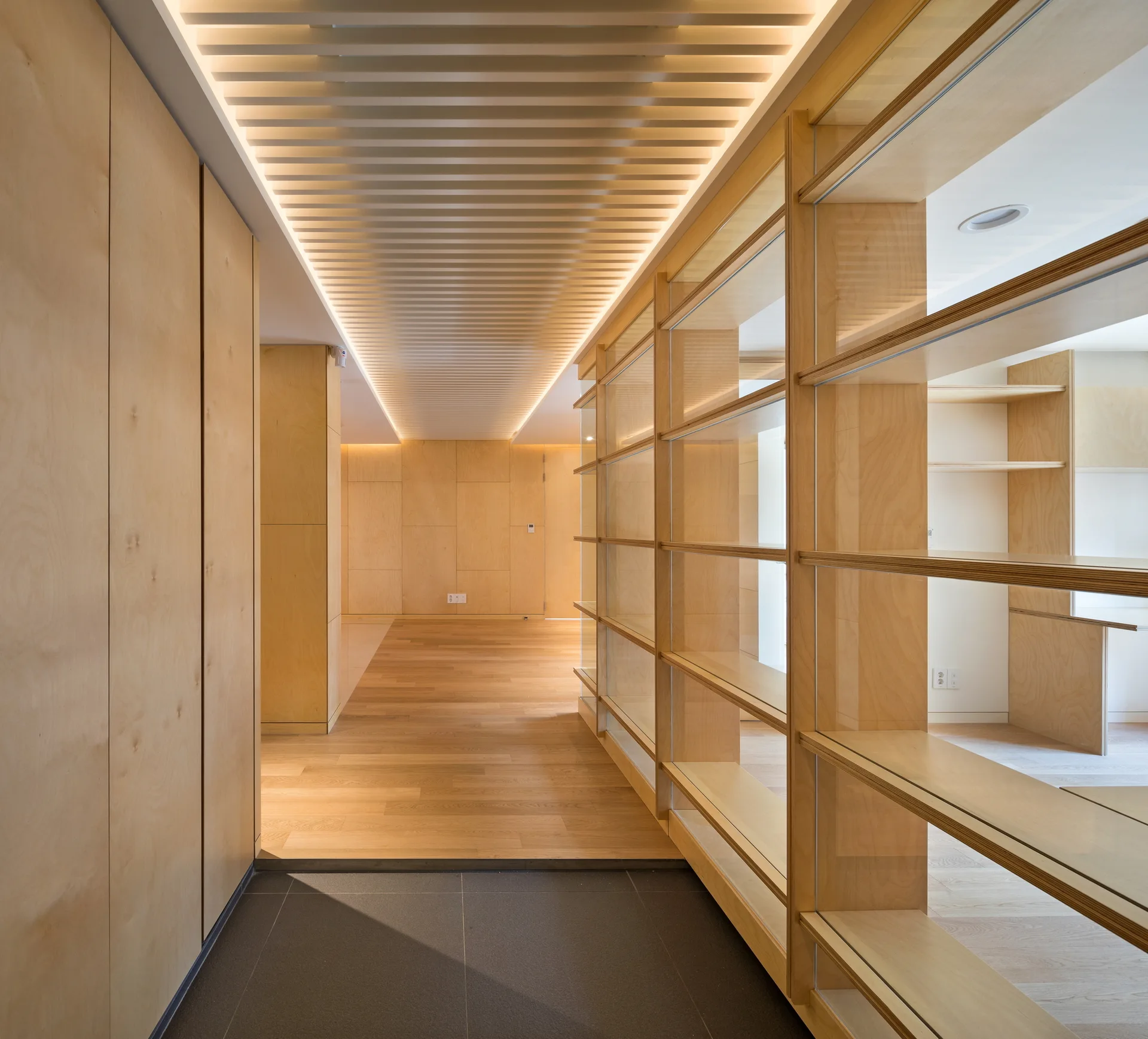 A minimalist hallway lined with light wood cabinets and glass-backed shelves features a linear slatted ceiling illuminated by hidden cove lighting.