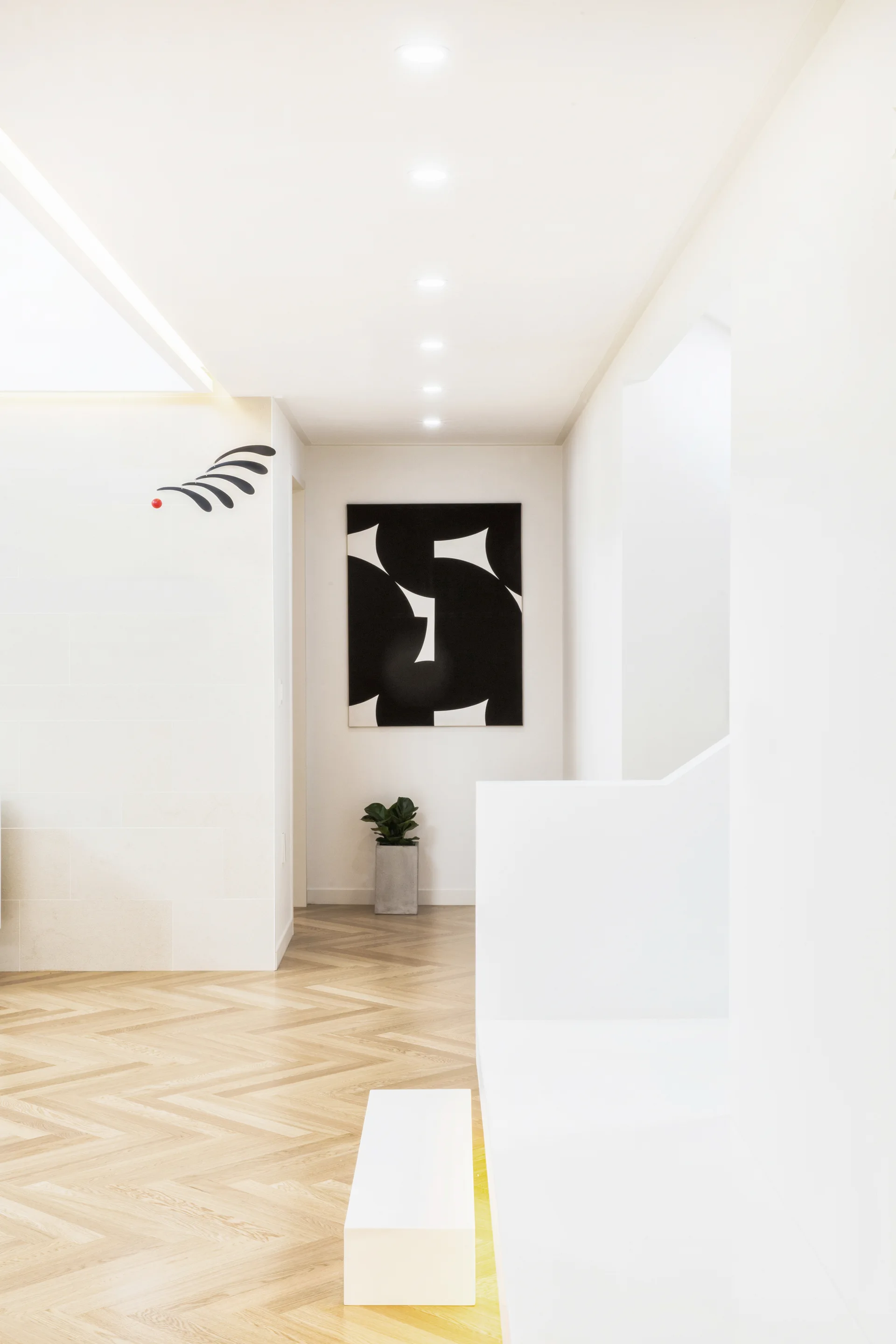 A minimalist white hallway features herringbone pattern wood flooring, a row of recessed ceiling lights, and a large black and white abstract painting.