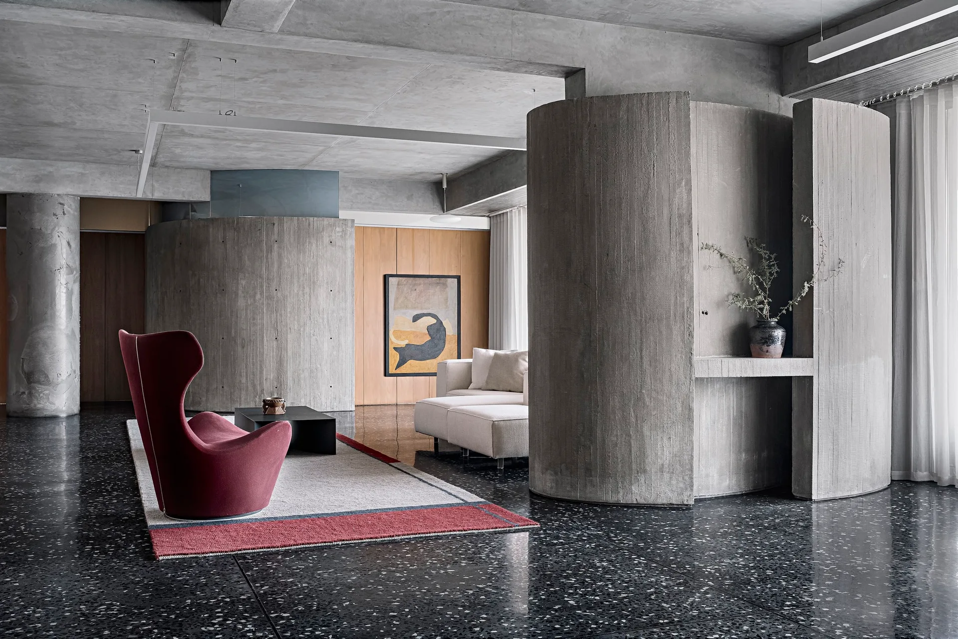 A minimalist living space features curved raw concrete partitions, a dark polished terrazzo floor, and a sculptural burgundy lounge chair on an area rug.