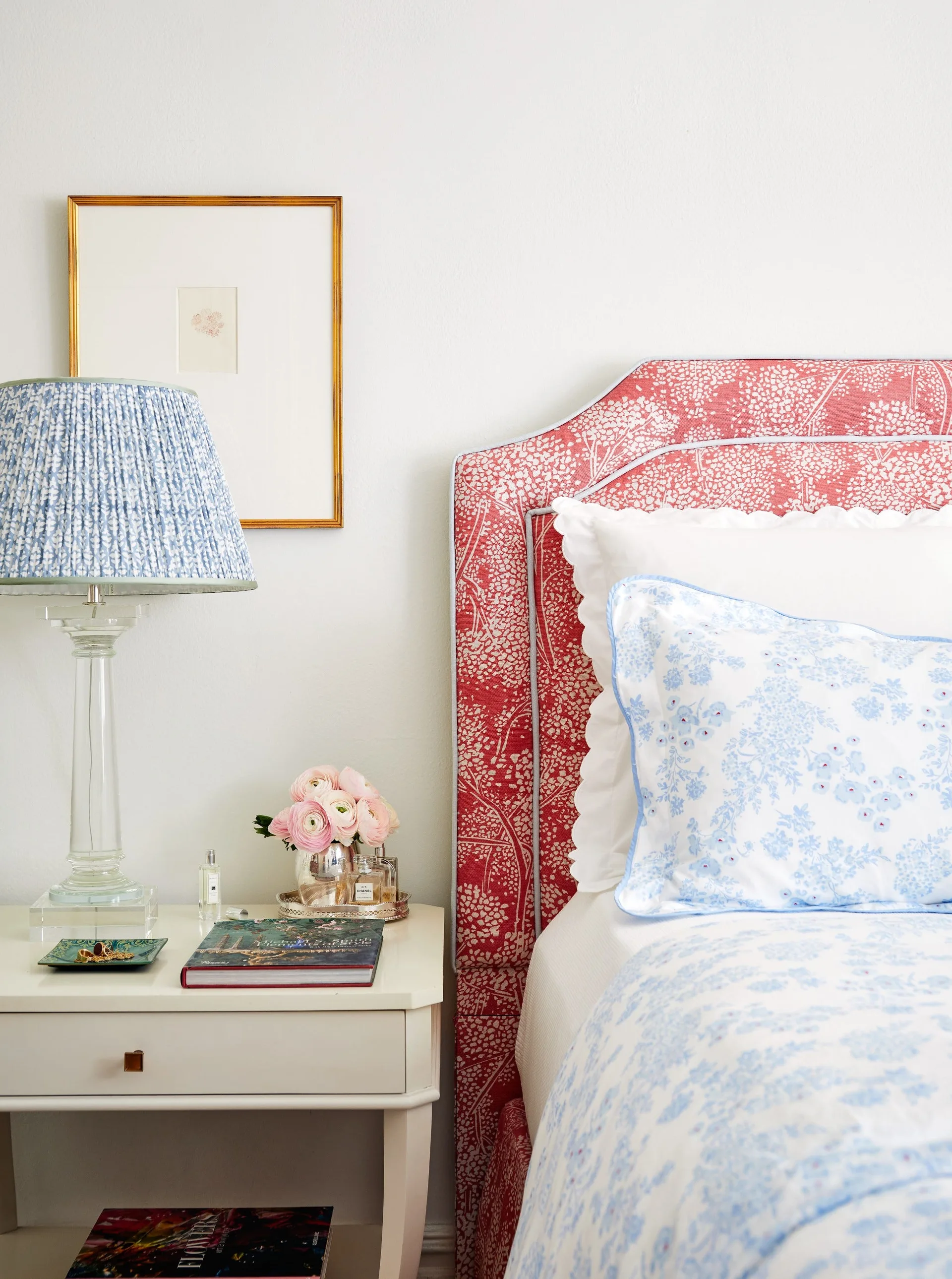 A bedroom corner features a red and white floral upholstered headboard next to a white nightstand topped with a blue pleated-shade lamp.